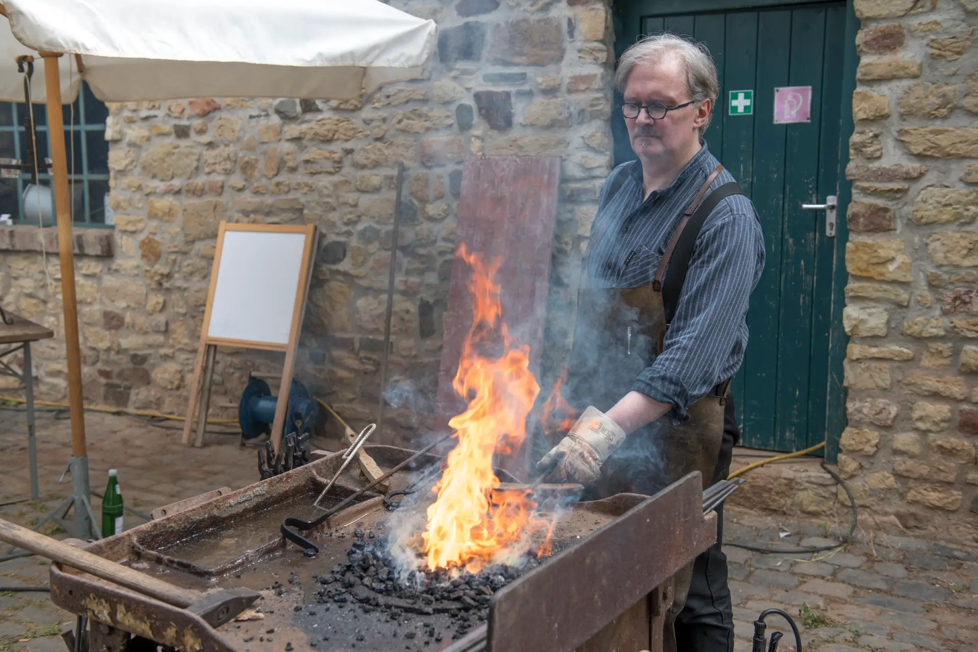 Ein Mann steht vor einem Feuer, das auf einem Metalltisch entzündet ist. Er trägt Handschuhe und hält eine Eisenstange ins Feuer. Hinter ihm ist eine grüne Tür und die Mauer eines Gebäudes.