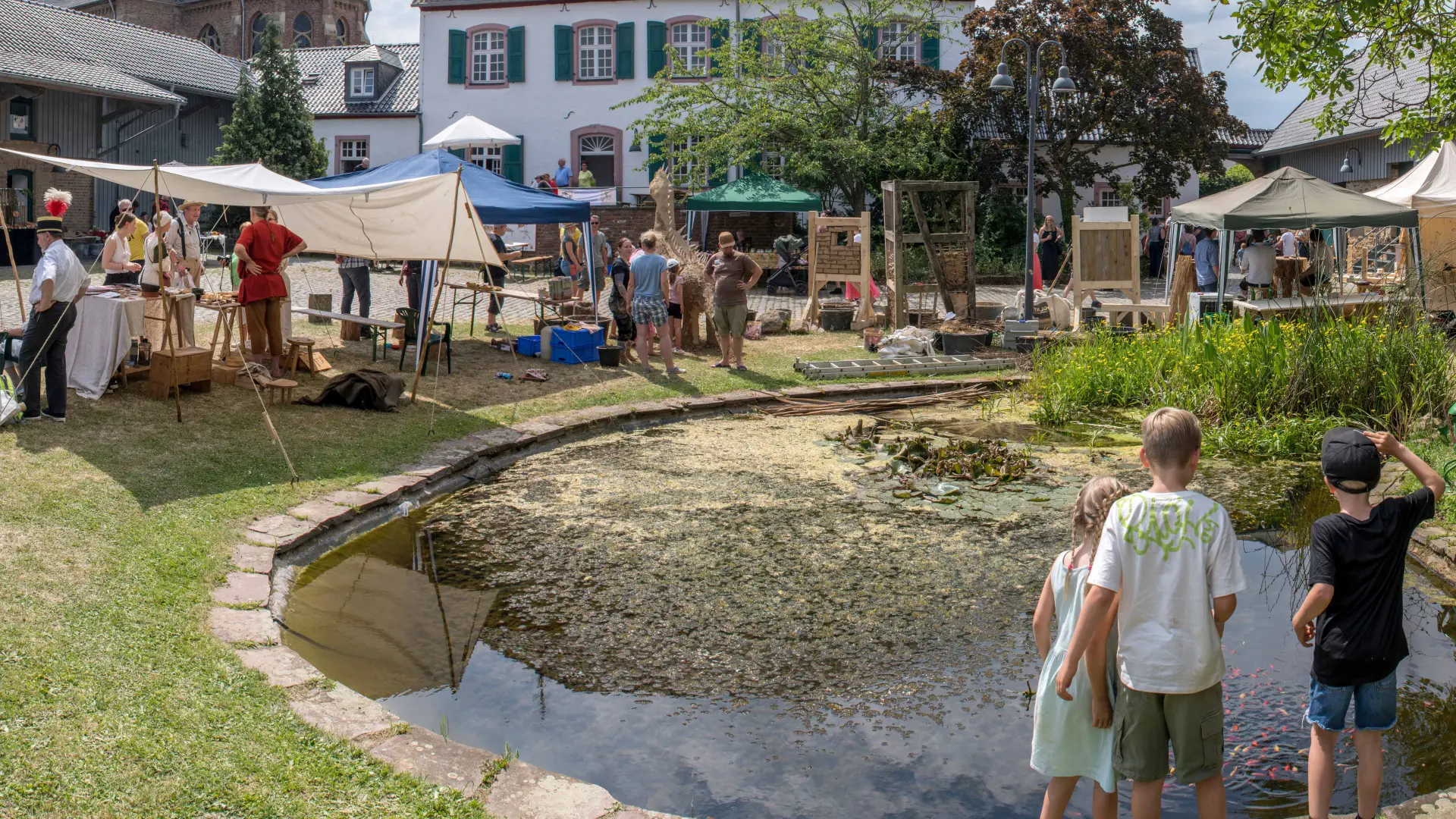 Übersichtsfoto einer Veranstaltung auf dem Stiftshof mit einigen Ständen an denen archäologiebezogene Themen vorstellen. Im Vordergrund sind drei Kinder, die an dem kleinen Teich stehen und daraufblicken.