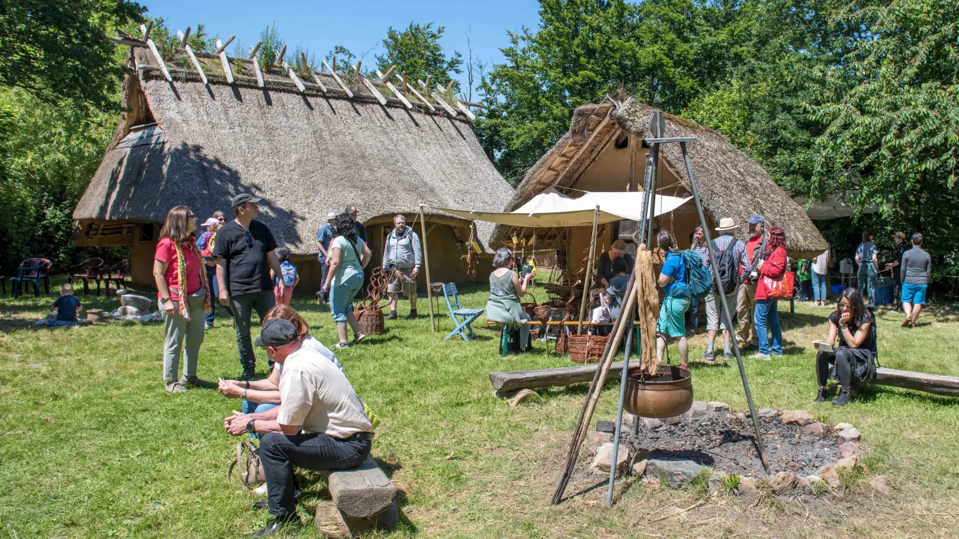 Zwei nachgebaute eisenzeitliche Häuser mit Reetdach auf einer Wiese. Einige Personen laufen durch das Bild oder sitzen auf Bänken vor dem Haus.