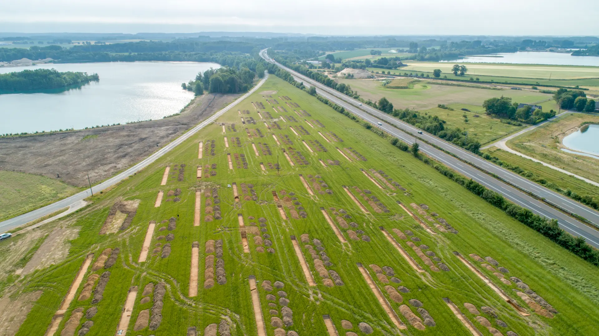 Foto einer grünen Wiese aus der Luft, auf der mehrere schmale, langrechteckige Grabungsschnitte zu sehen sind, im Hintergrund drei Seen und Wälder am Horizont.