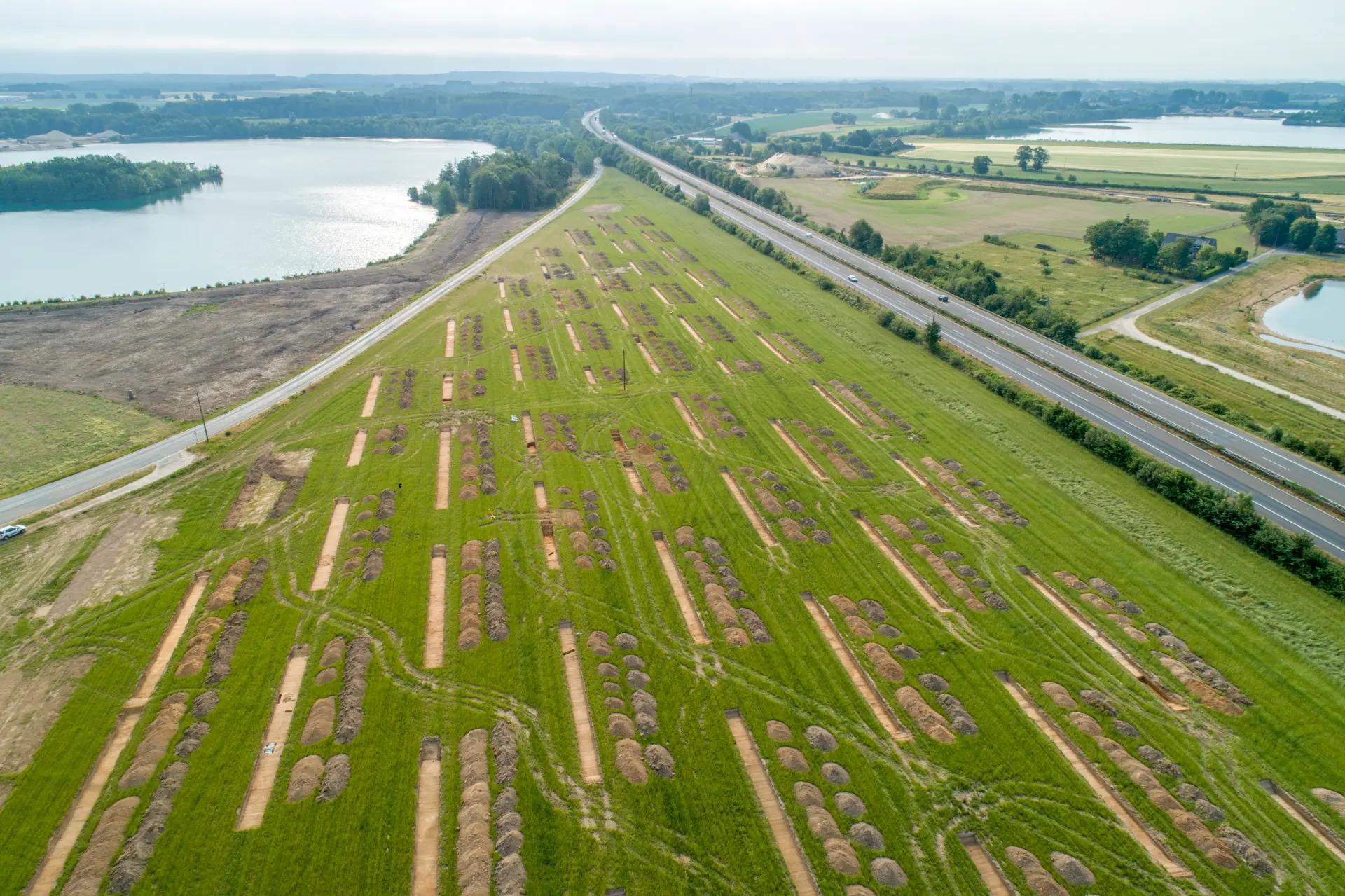 Foto einer grünen Wiese aus der Luft, auf der mehrere schmale, langrechteckige Grabungsschnitte zu sehen sind, im Hintergrund drei Seen und Wälder am Horizont.