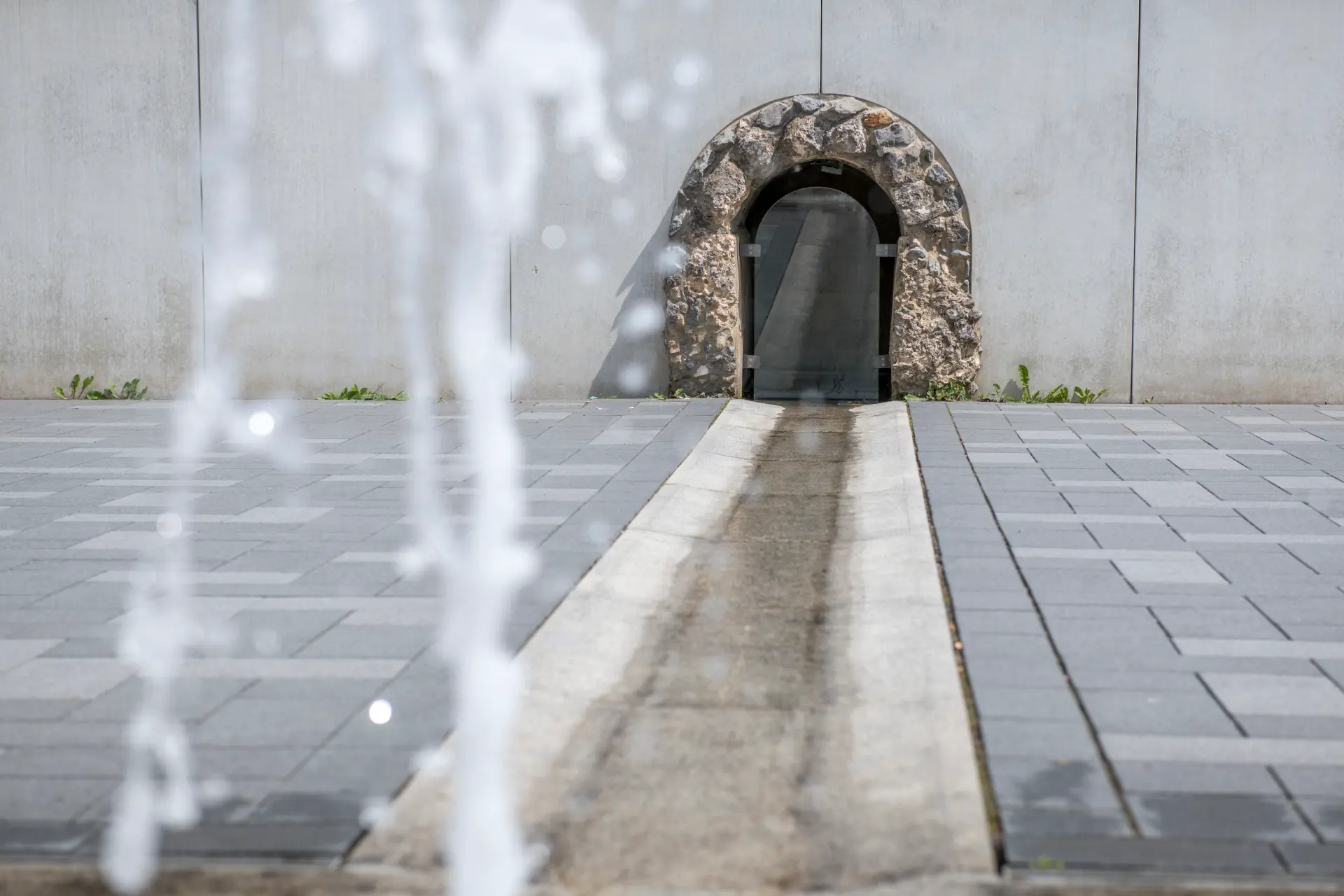 Blick über einen grau gepflasterten Platz hinweg auf einen tunnelförmigen Kanal, der in einer grauen Mauer eingelassen ist, im Vordergrund ist unscharf eine hochspritzende Wasserfontäne zu sehen.