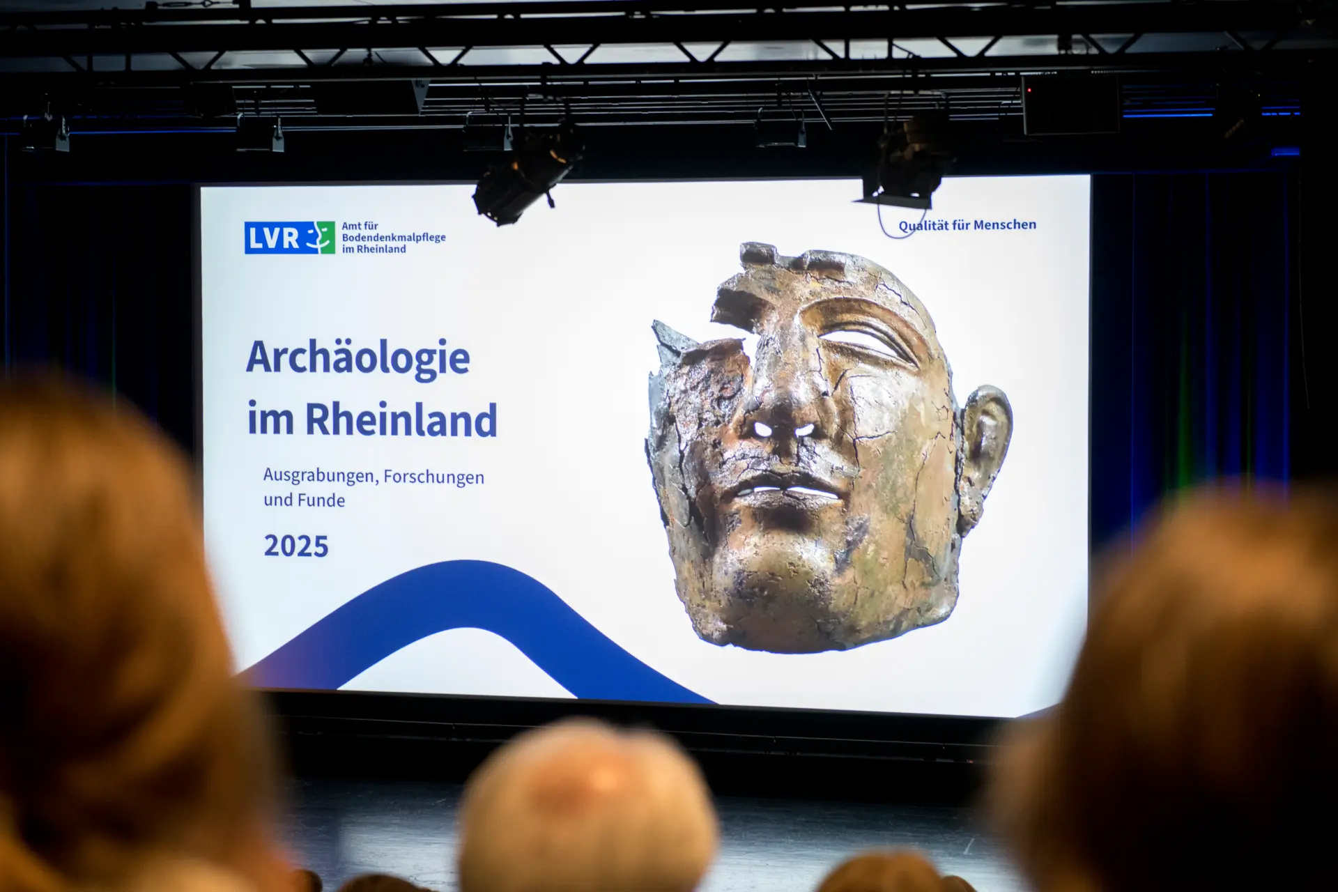 Blick auf eine per Beamer auf eine Leinwand projizierte Präsentationsfolie auf einer Bühne, im Vordergrund sind unscharf Hinterköpfe zu sehen.