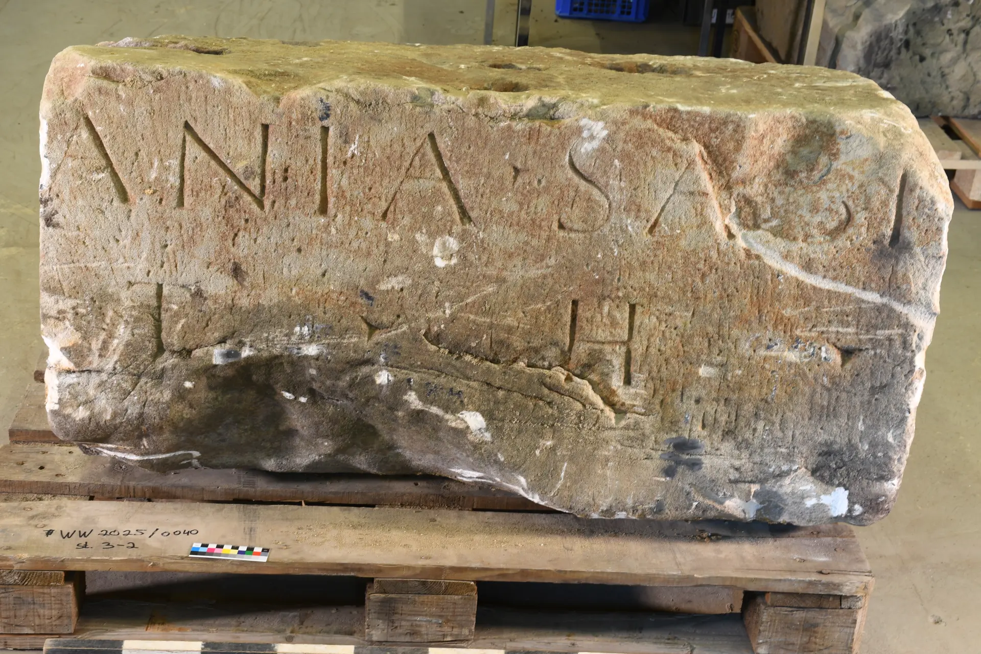Ein massiver grauer und leicht beschädigter Steinblock liegt auf einer Holzpalette, auf dem Block sind eingemeißelte Buchstaben zu erkennen.