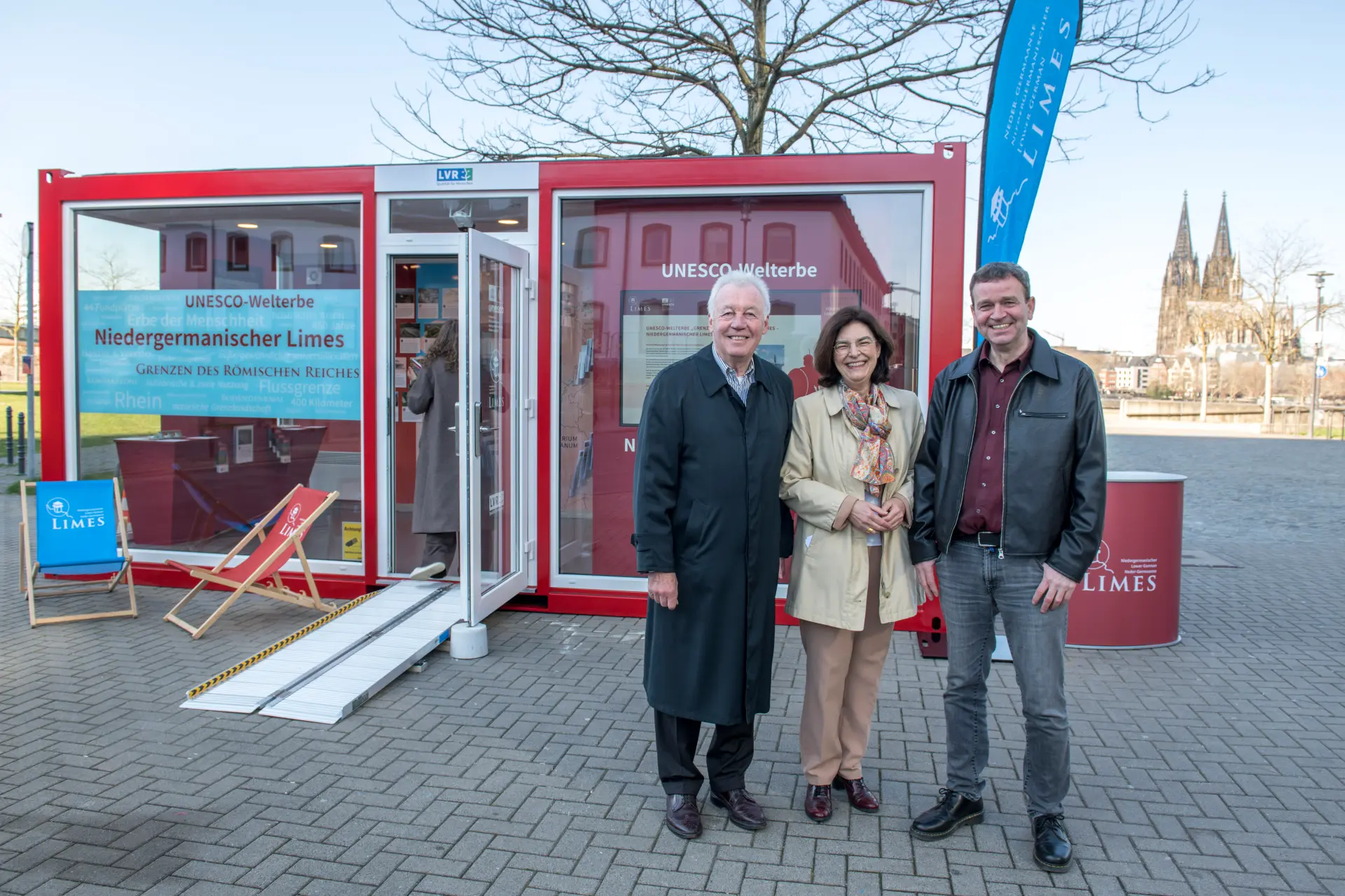 Drei Personen stehen vor einem roten Glascontainer und lächeln in die Kamera, im Hintergrund ist in der Ferne der Kölner Dom zu erkennen.