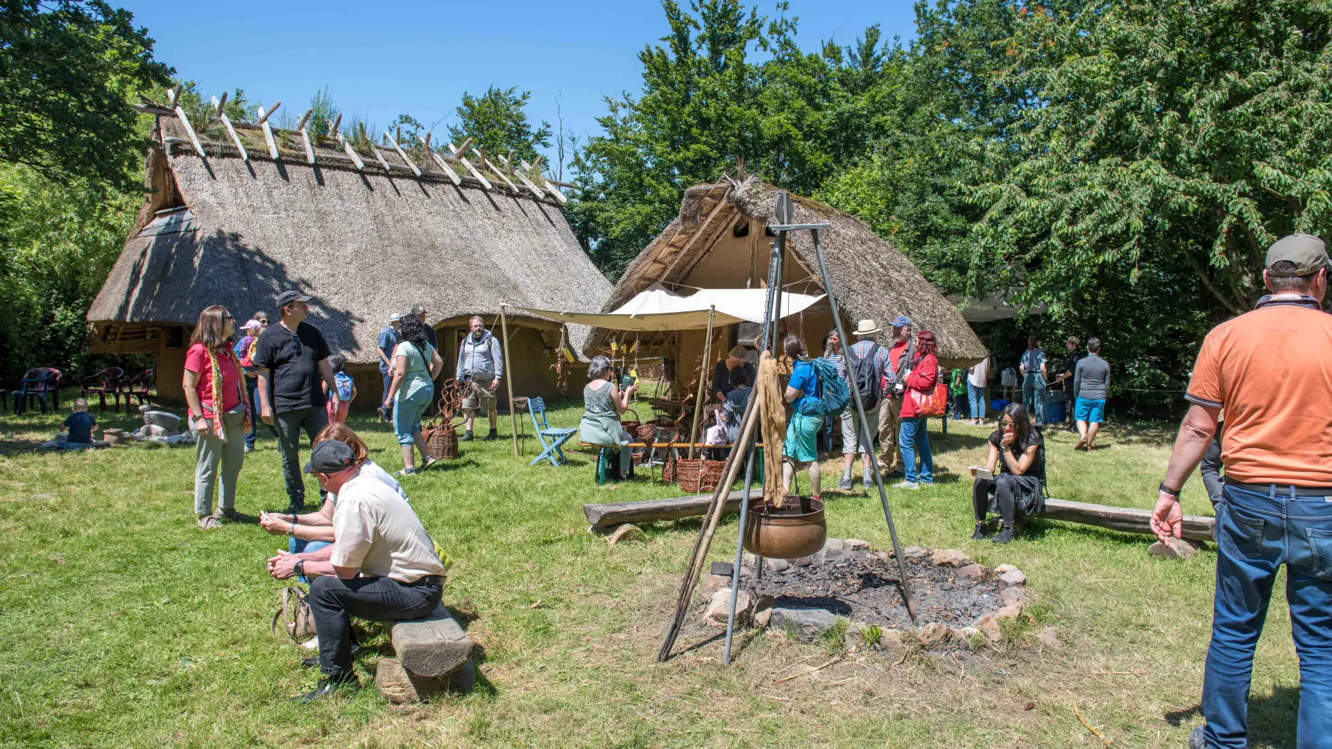 Zwei nachgebaute eisenzeitliche Häuser mit Reetdach auf einer Wiese. Einige Personen laufen durch das Bild oder sitzen auf Bänken vor dem Haus.