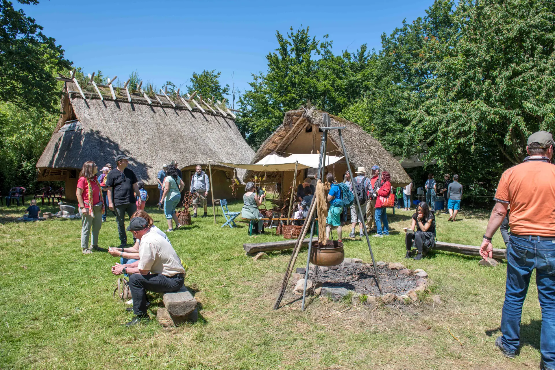 Zwei nachgebaute eisenzeitliche Häuser mit Reetdach auf einer Wiese. Einige Personen laufen durch das Bild oder sitzen auf Bänken vor dem Haus.