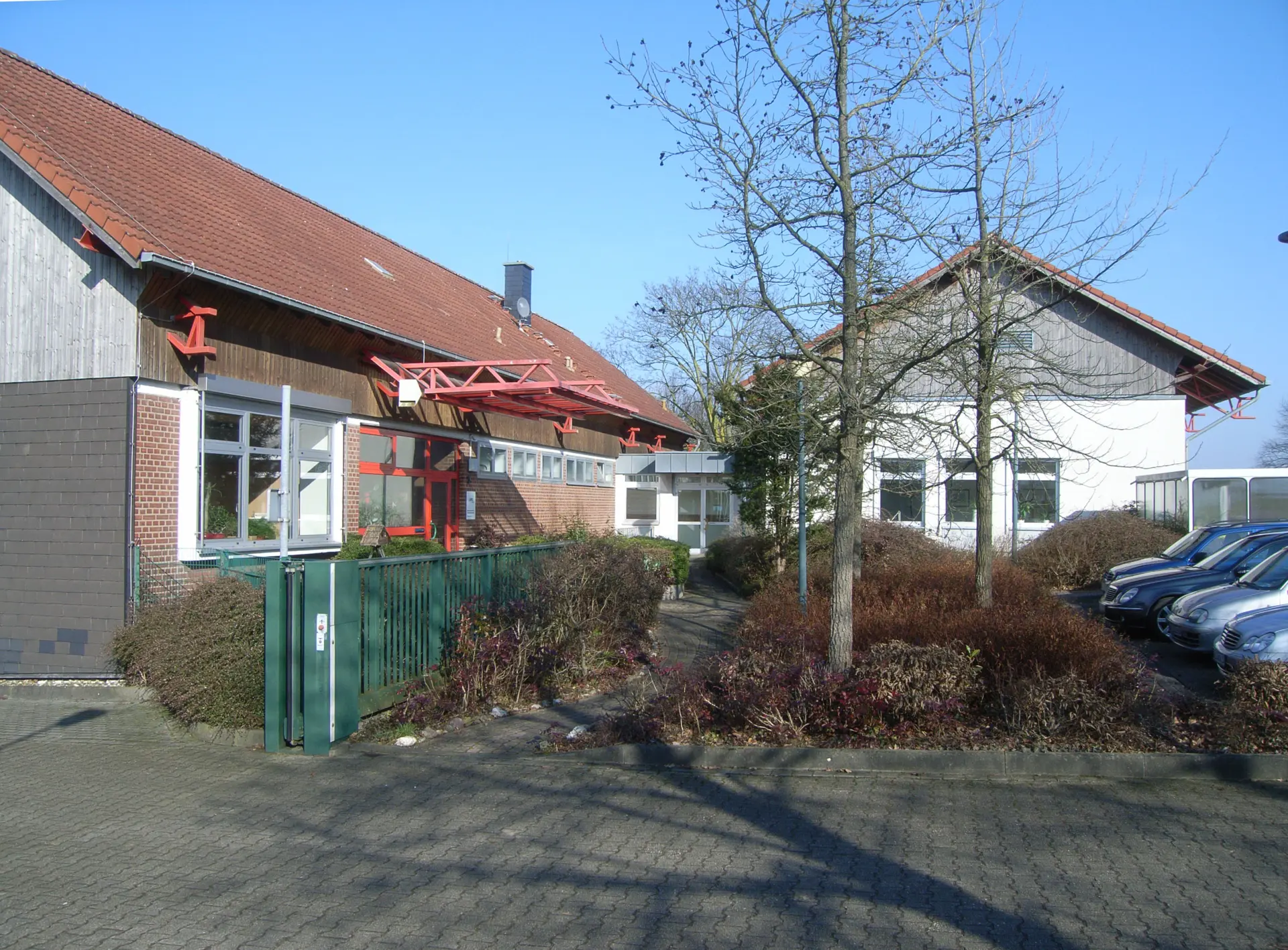 Der Eingangsbereich eines Gebäudekomplexes. Ein kleiner weg führt zur Tür entlang. Neben dem Weg wachsen Büsche. Rechts ist ein Teil des Parkplatzes mit Autos zu sehen.