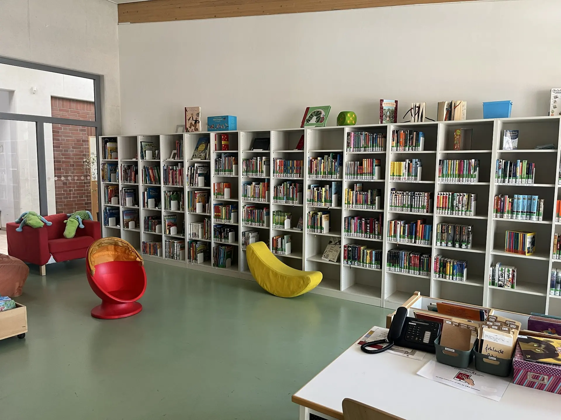 Freundlicher Raum mit Sitzgelegenheiten und einem langen Regal mit vielen Büchern an der der Wand