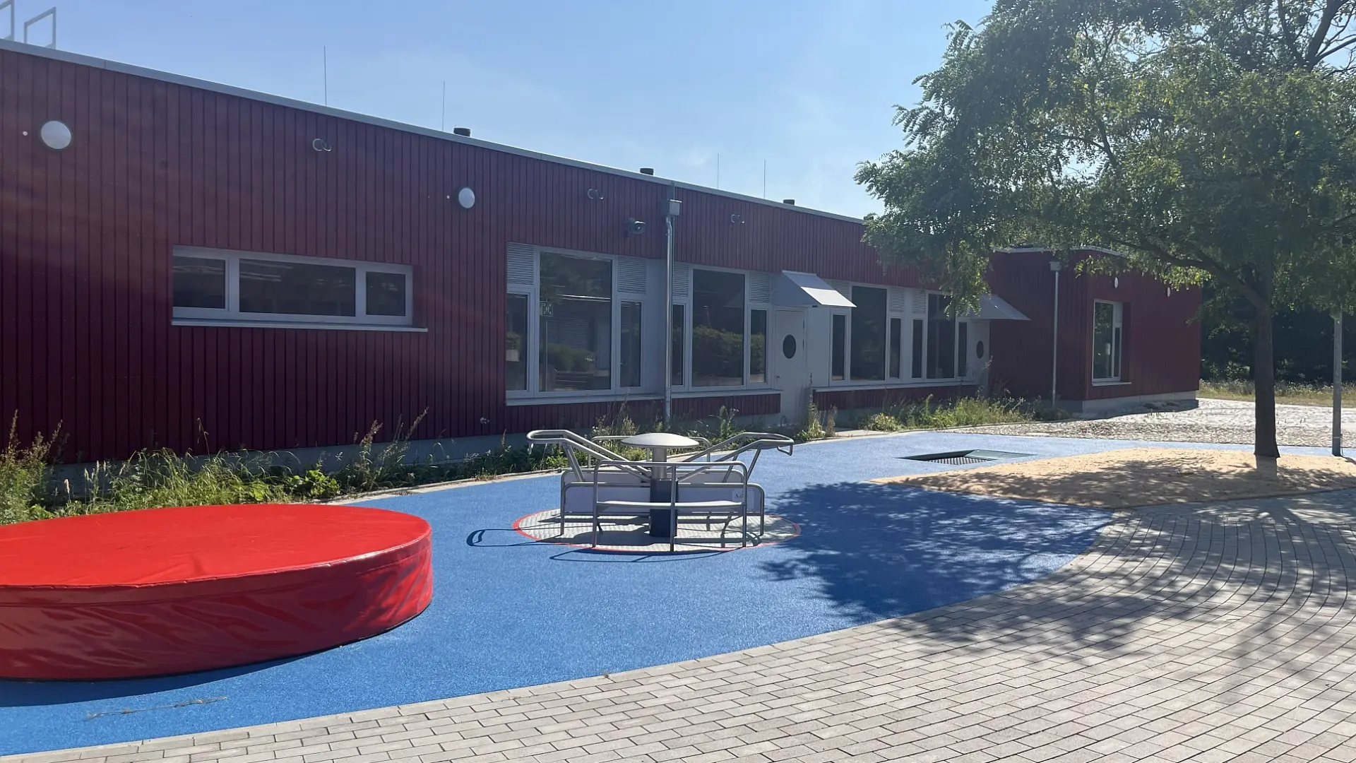 Spielplatz mit Karussell, rotem Trampolin und blauem Boden vor Schule