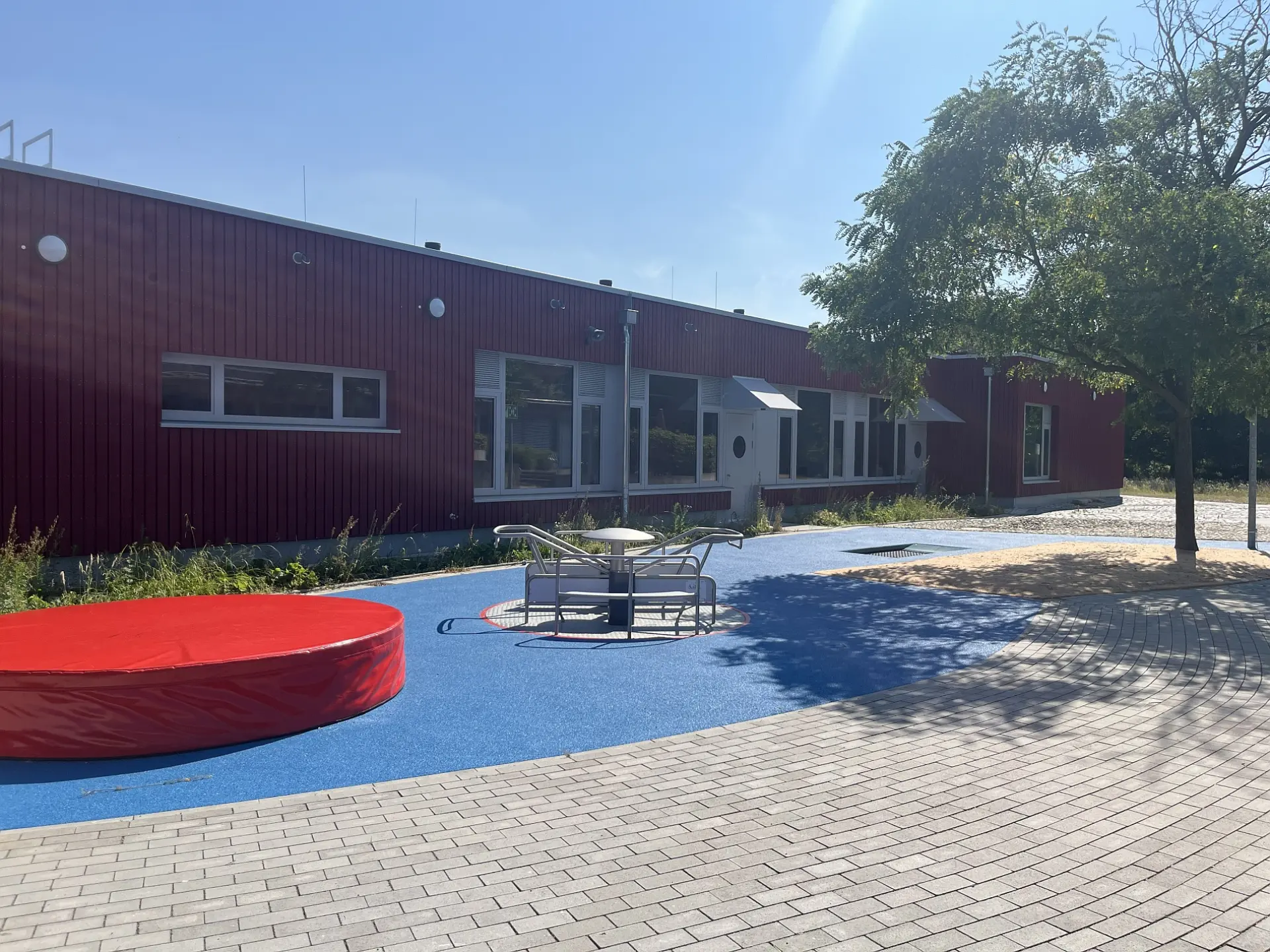 Spielplatz mit Karussell, rotem Trampolin und blauem Boden vor Schule