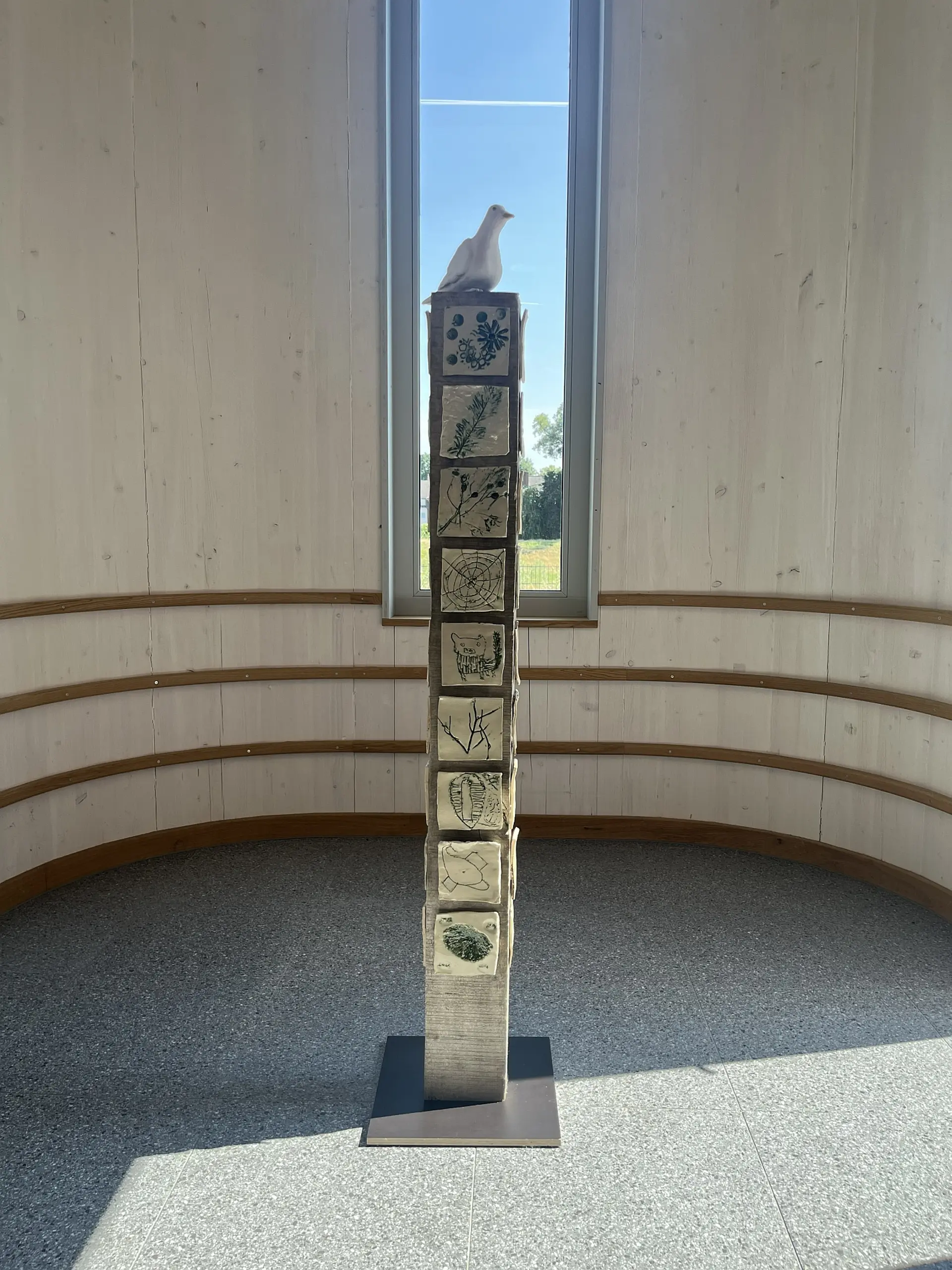Gedenkstele mit verzierten Ton-Fliesen und Friedenstaube vor Fenster