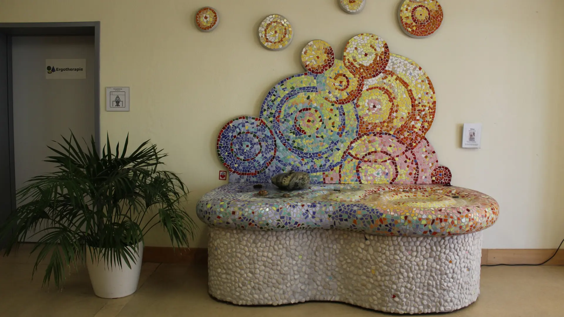Der Erinnerungsbrunnen als eine Sitzbank-ähnliche Konstruktion aus Stein. Sie ist zahlreichen bunten Mosaiksteinen bedeckt.