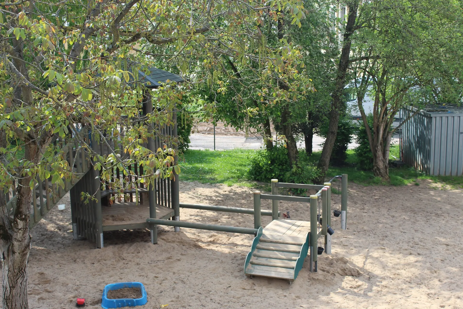 Ein Teil des Schulhofs mit einem Holzklettergerüst, viel Sand auf dem Boden und vielen grünen Bäumen drum-herum