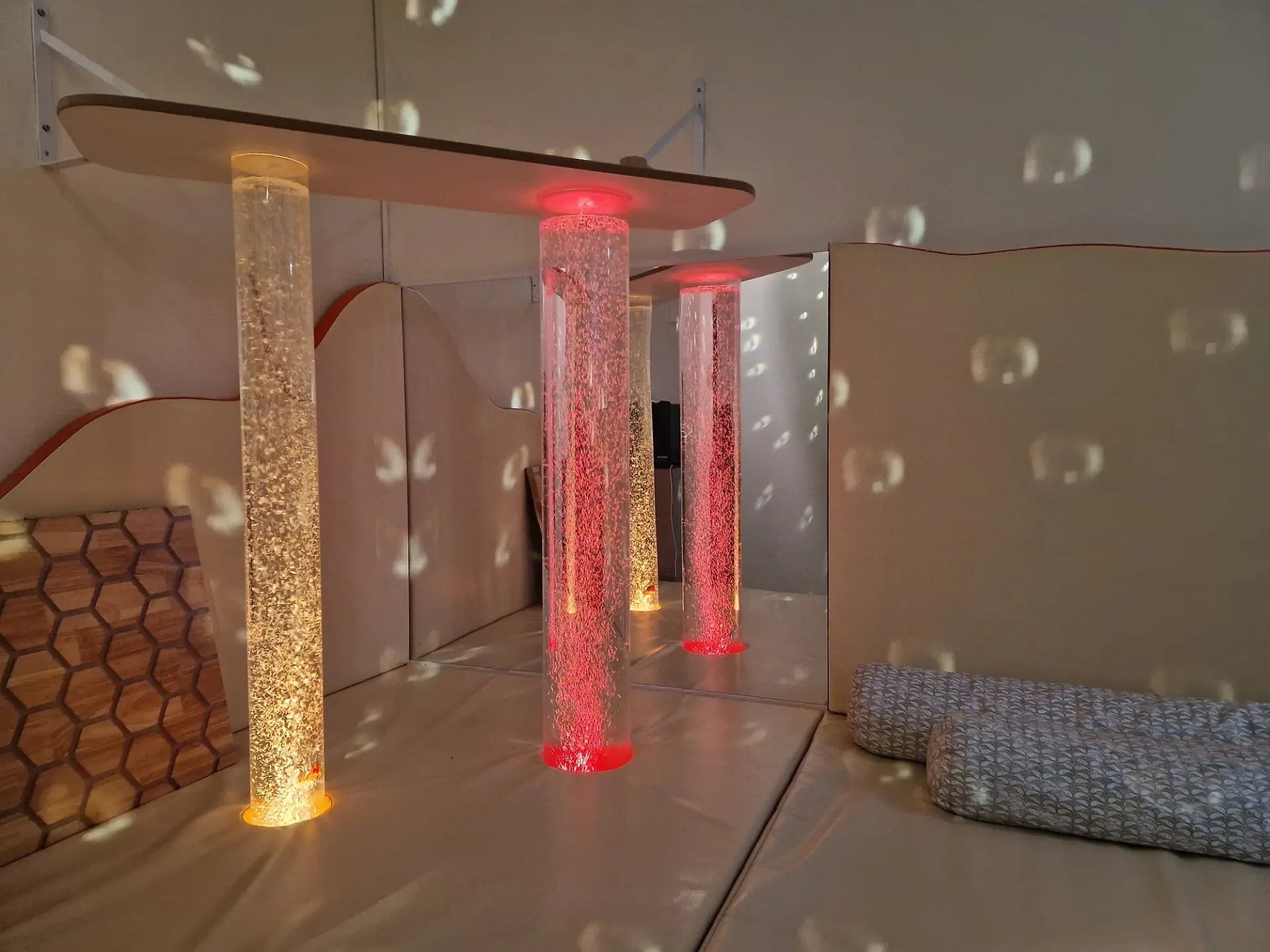 Ein Snoezelraum mit großen Kissen auf dem Boden, einer Licht-Berührungstafel und drei großen Säulen, die voll mit Wasserblasen sind und in verschiedenen Farben leuchten.