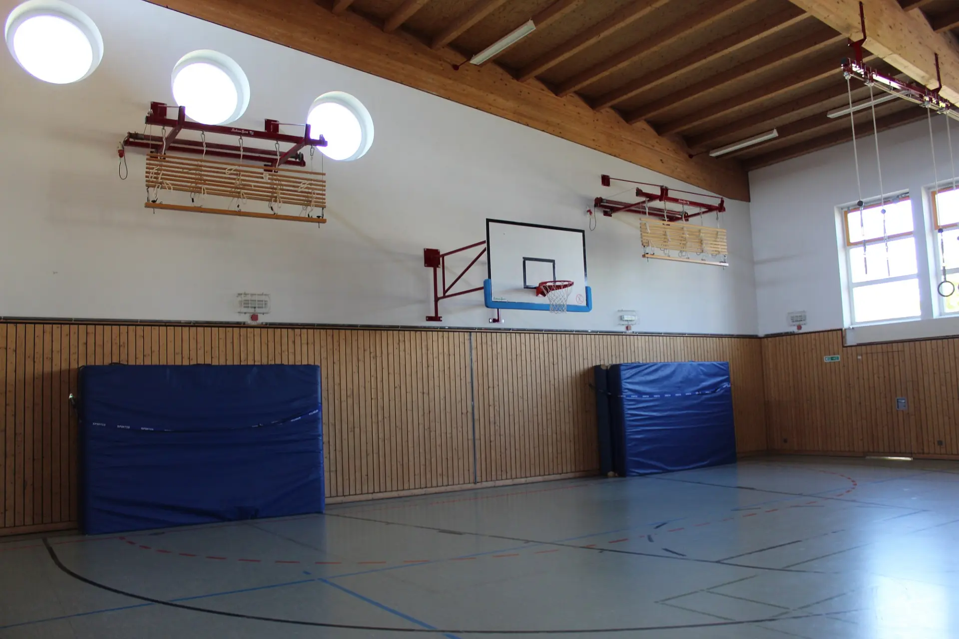Die linke Hälfte einer modern ausgestatteten Sporthalle mit Sprossenwänden, Basketballkorb und Ringen.