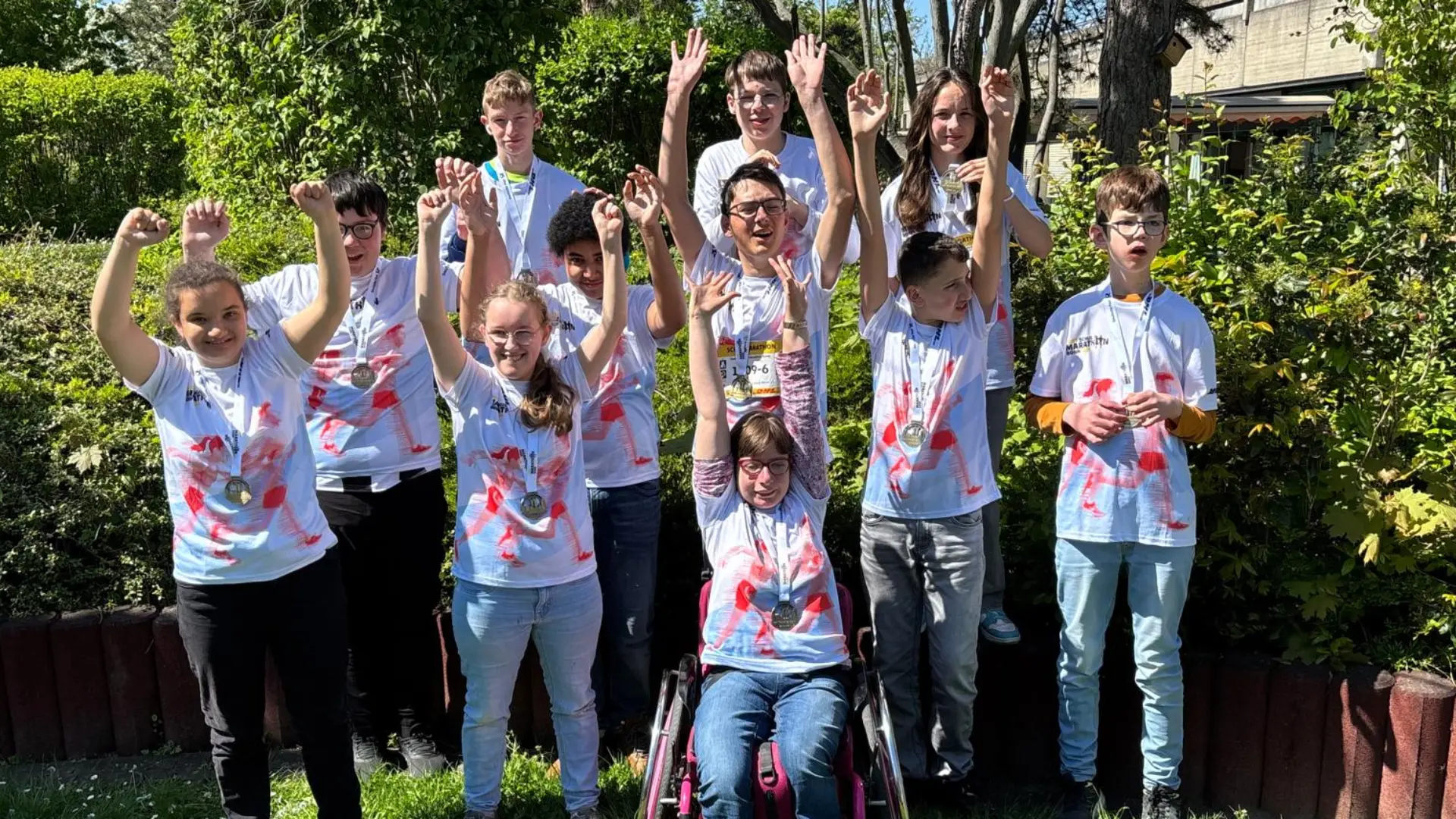 Elf Schüler*innen, alle mit Bonner Marathon-T-Shirts und Medaillen, lächeln und halten ihre Arme jubelnd in die Luft.