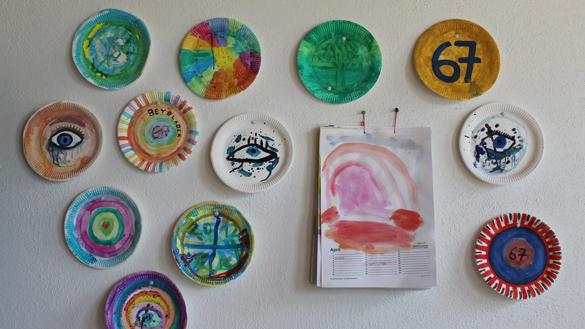 Eine weiße Wand, an der mehrere bunt bemalte Pappteller und ein aufgeschlagener Wandkalender mit einer farbigen Zeichnung hängen.