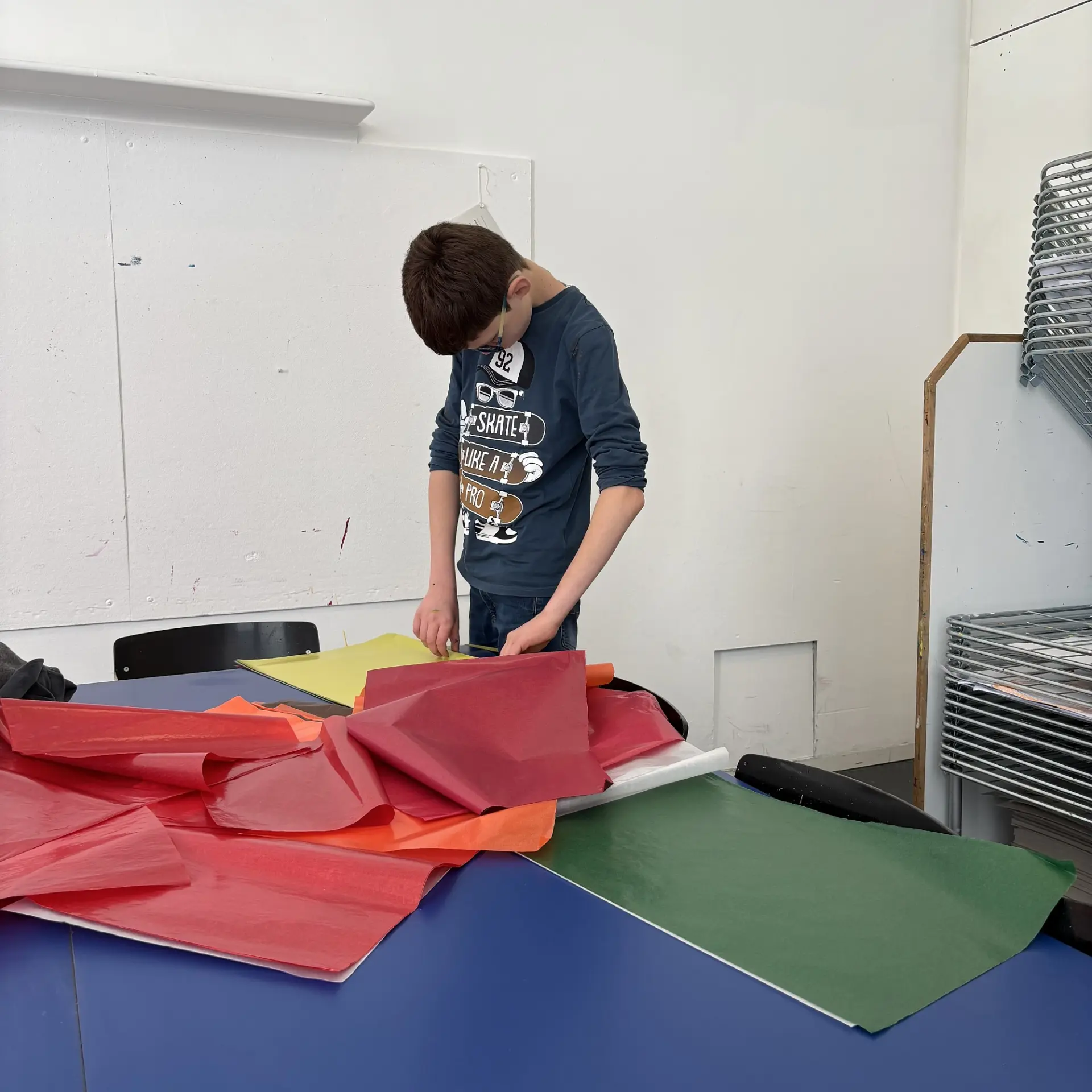 Ein Junge steht an einem großen Tisch und blickt konzentriert nach unten auf ein Stück gelbes Papier, das vor ihm liegt.
