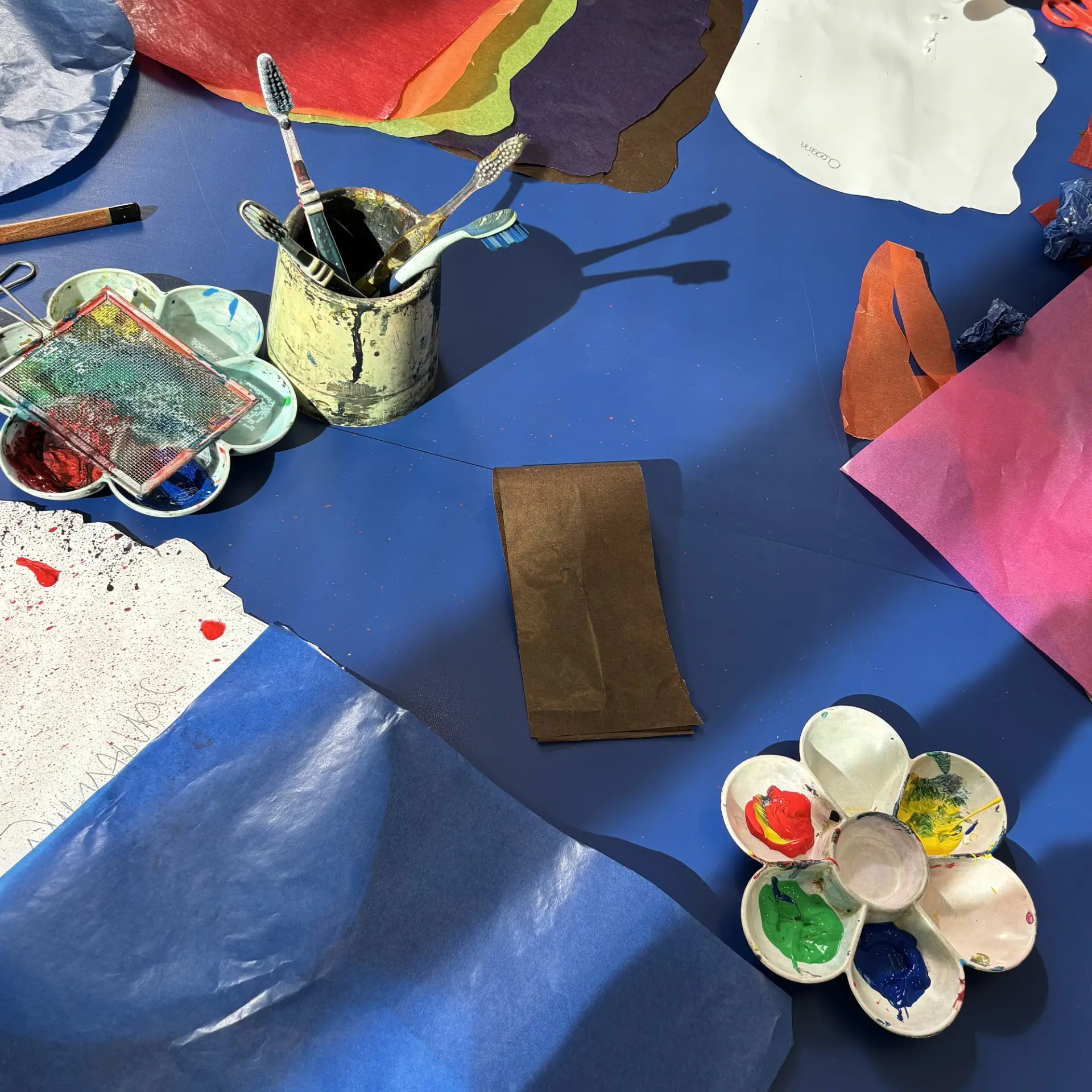Malutensilien, zwei Mischpaletten mit Farben, Zahnbürsten im Becher und buntes Papier auf blauem Tisch.