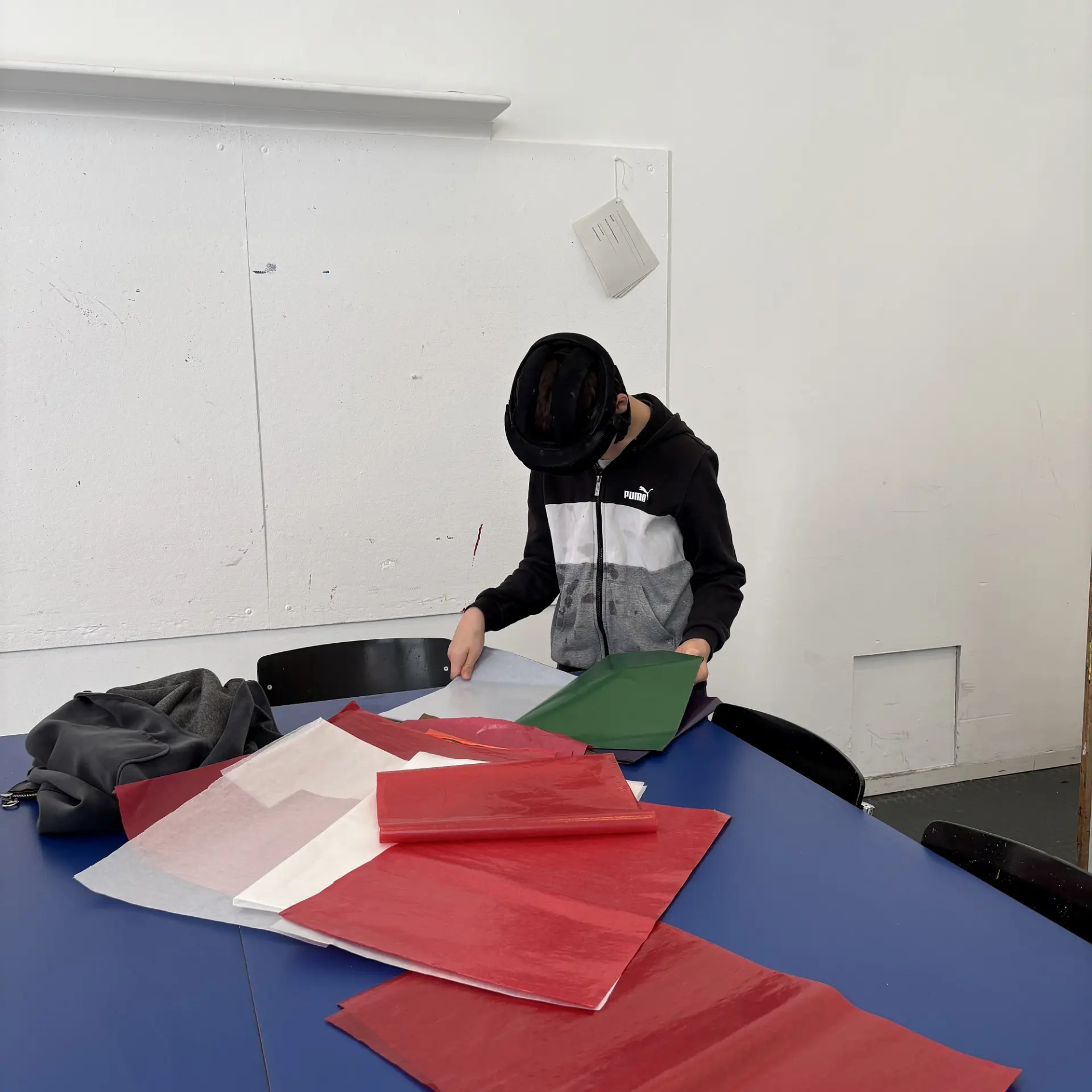 Ein Junge mit Kopfschutz steht an einem blauen Tisch und sortiert rotes, weißes und grünes Papier.