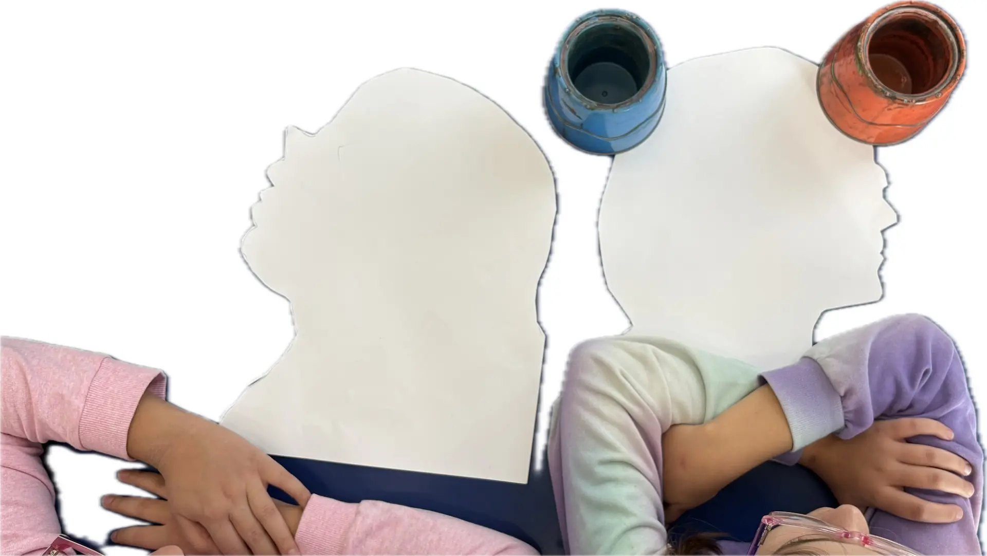 Draufsicht auf einen Tisch, an dem zwei Kinder sitzen. Vor ihnen liegt jeweils eine große Silhouette eines Kopfprofils aus Papier.