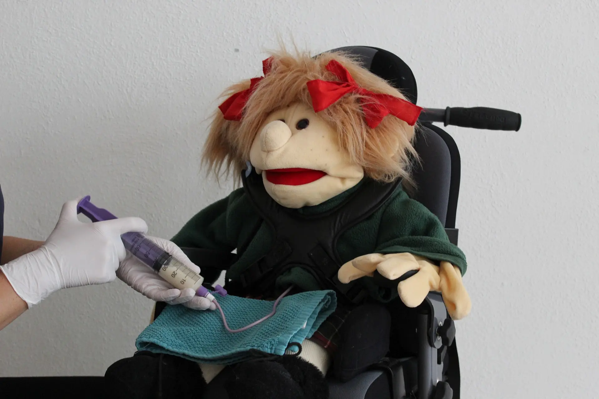 Die Hände einer Person, die einer Stoffpuppe im Rollstuhl Nahrung über einen Katheter gibt.