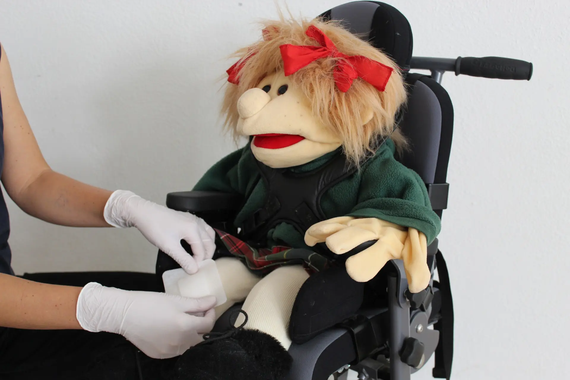 Die Hände einer Person, die einen Pflaster ans Knie einer Stoffpuppe im Rollstuhl anbringt.