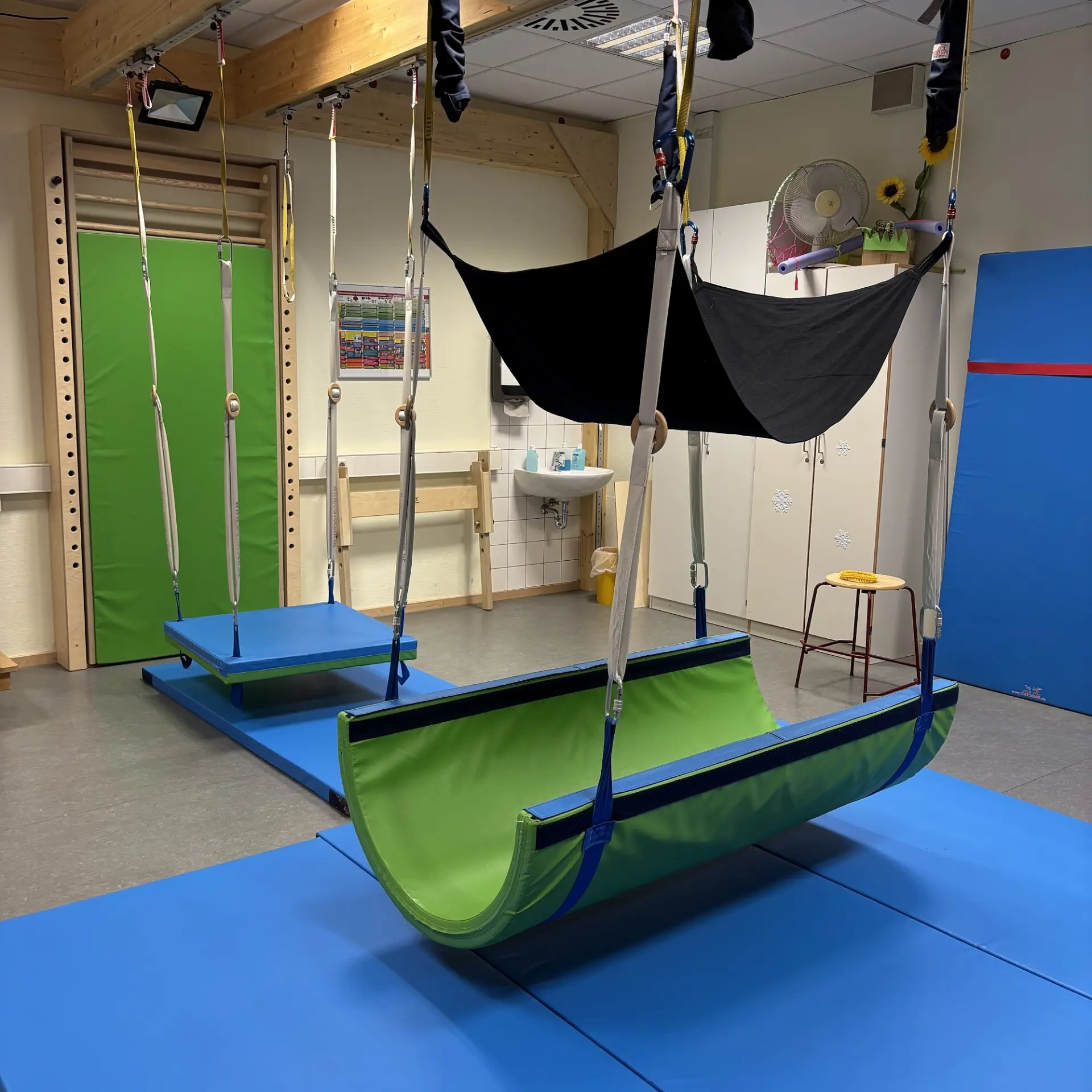 Ein Therapieraum mit verschiedenen Geräten, unter anderem drei Liege- und Sitzschaukeln, die von der Decke hängen.
