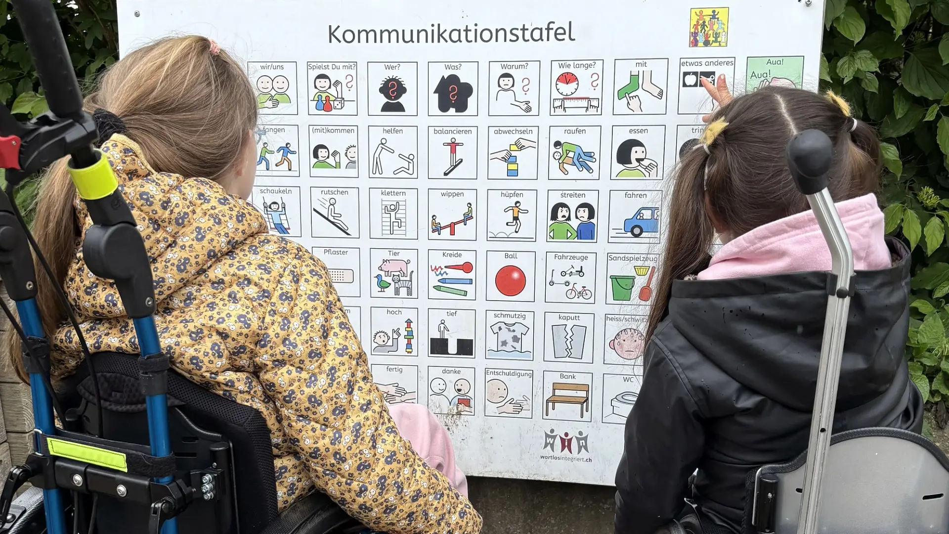 Zwei Schülerinnen im Rollstuhl stehen vor der Kommunikationstafel und schauen sich die Metacom-Symbole darauf an.