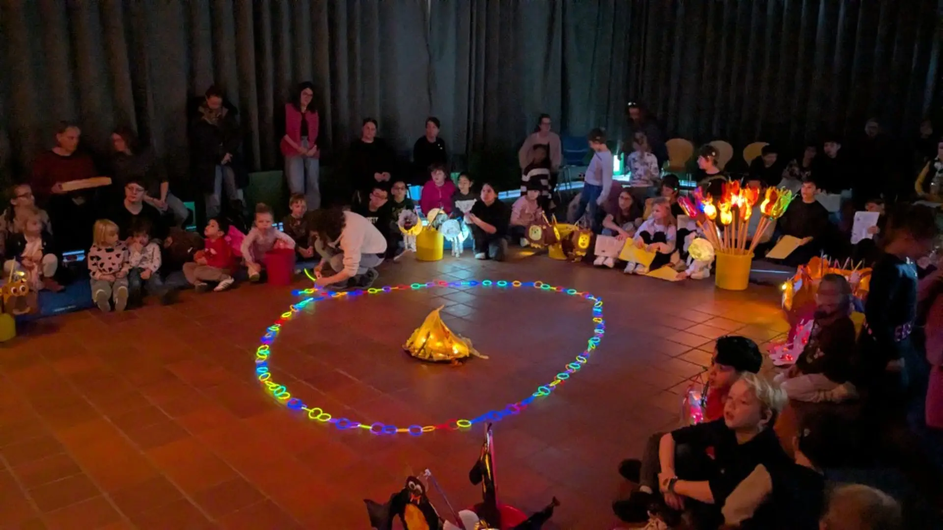 Blick in einen abgedunkelten Raum. Auf dem Boden sitzen Kinder in einem Halbkreis. In der Mitte ist ein Kreis aus einer Leuchtkette gelegt, der ein LED-Lagerfeuer umgibt.