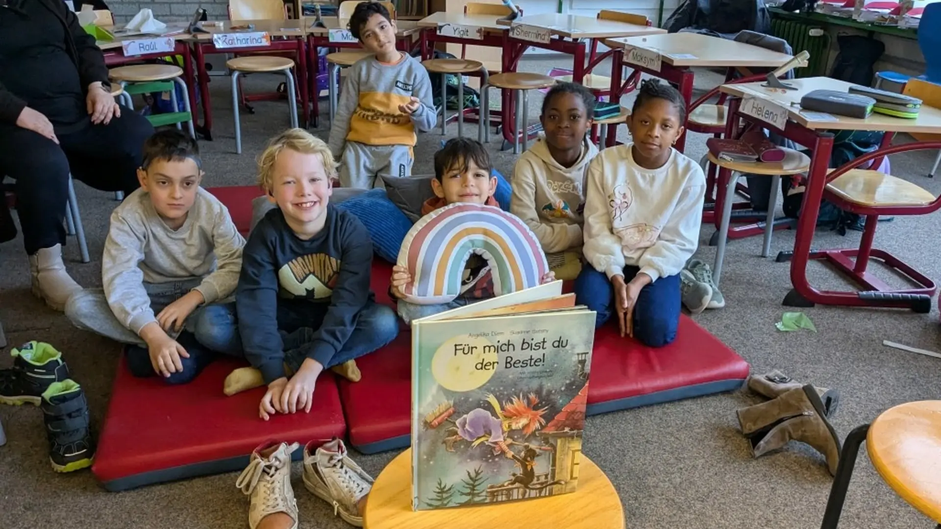 In einem Klassenraum sitzen Kinder auf einer Matte auf dem Fußboden. Ein Kind hält ein Regenbogen-Kissen in den Händen. Vor den Kindern steht ein großes Buch "Für mich bist du der Beste!"
