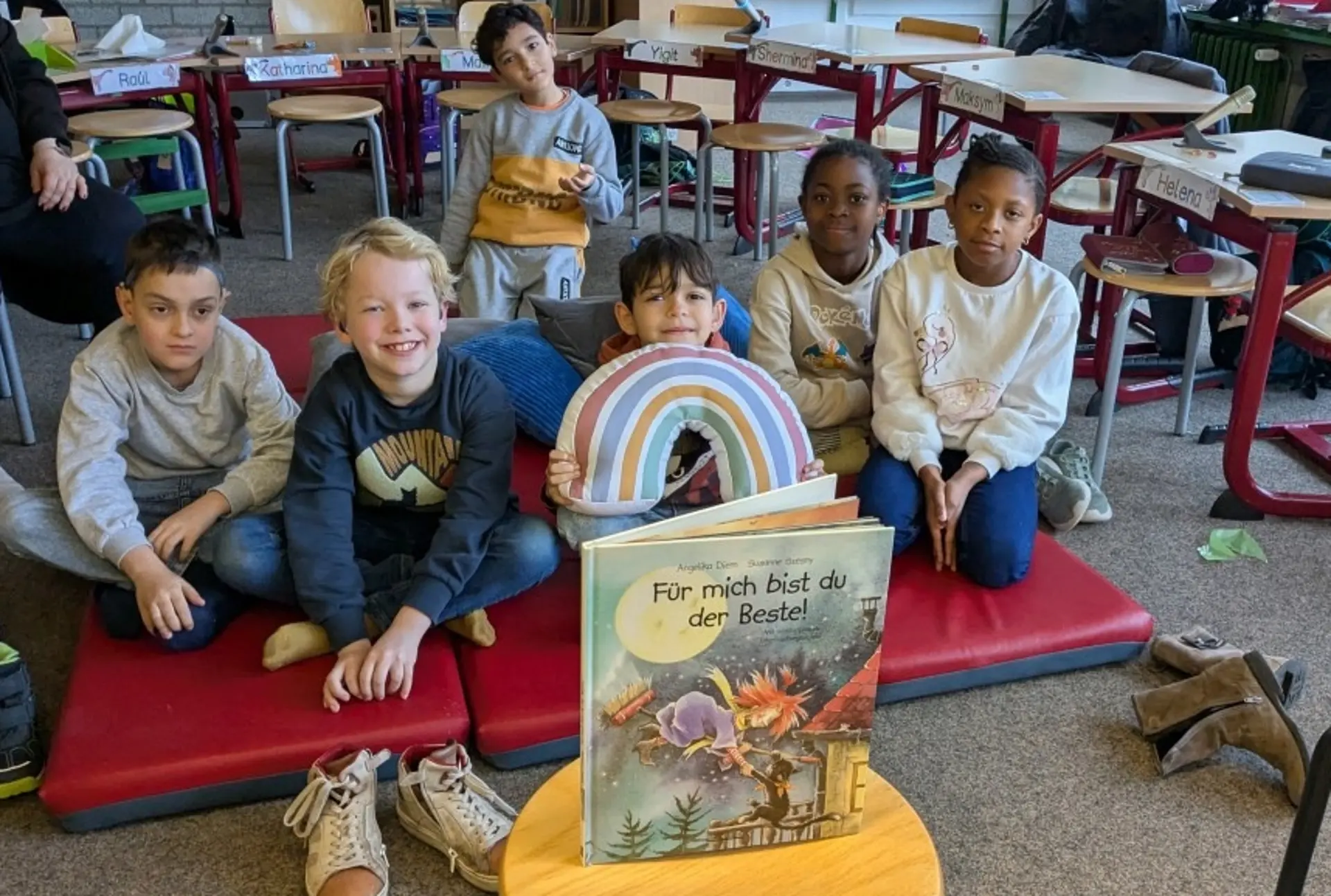 In einem Klassenraum sitzen Kinder auf einer Matte auf dem Fußboden. Ein Kind hält ein Regenbogen-Kissen in den Händen. Vor den Kindern steht ein großes Buch "Für mich bist du der Beste!"