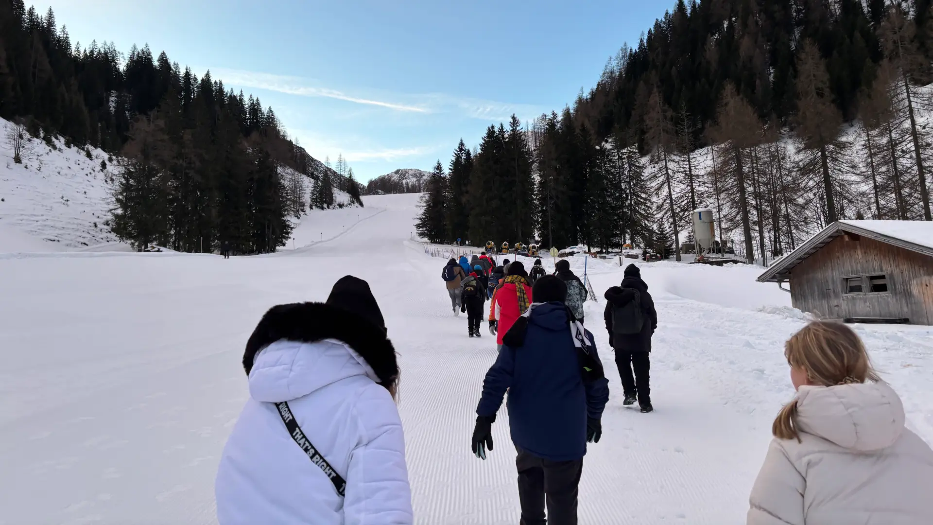 Eine Hand voll Menschen läuft eine flache Skipiste bei Tag hoch. Im Hintergrund ist eine kleine Hütte zu erkennen.