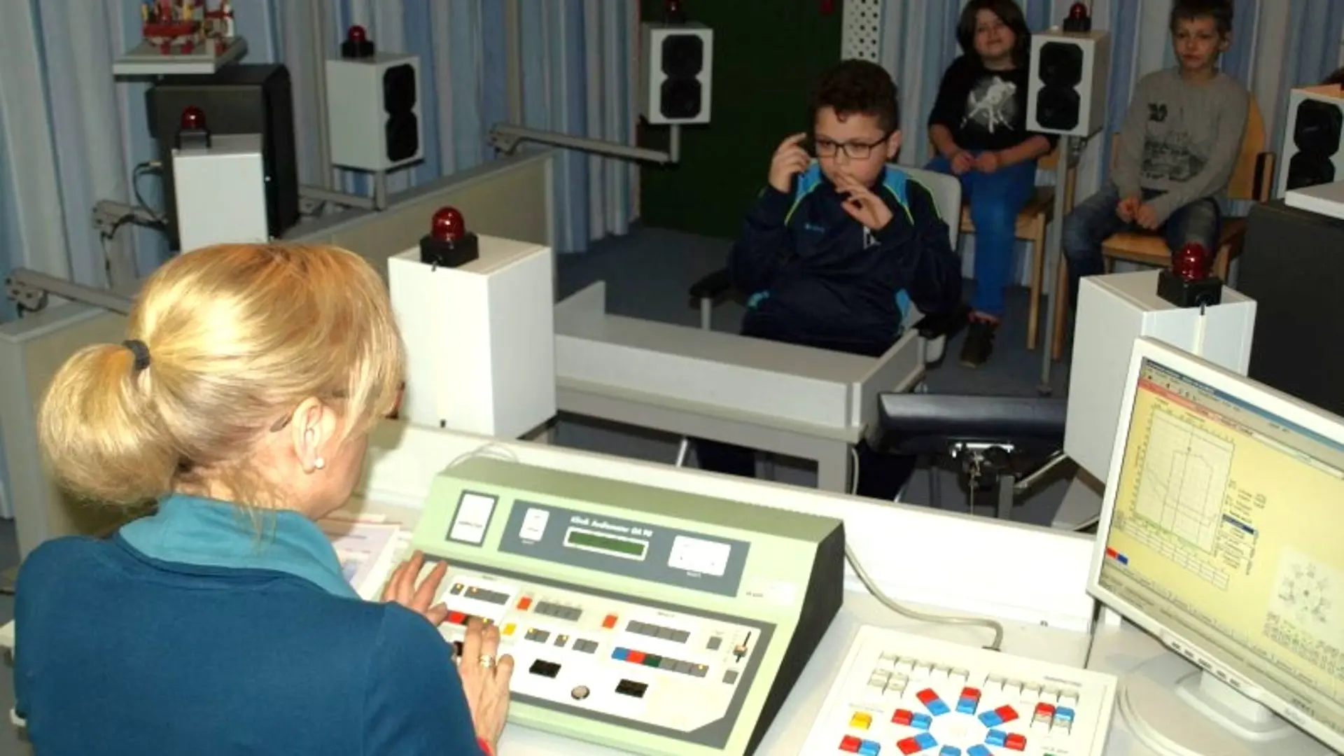 Eine audiometrische Untersuchung: Eine Frau spielt über ein technisches Gerät verschiede Töne über mehrere Lautsprecher aus. Vor ihr sitz ein Kind, das auf sein rechtes Ohr zeigt. Weiter hinten sitzen noch zwei Kinder.