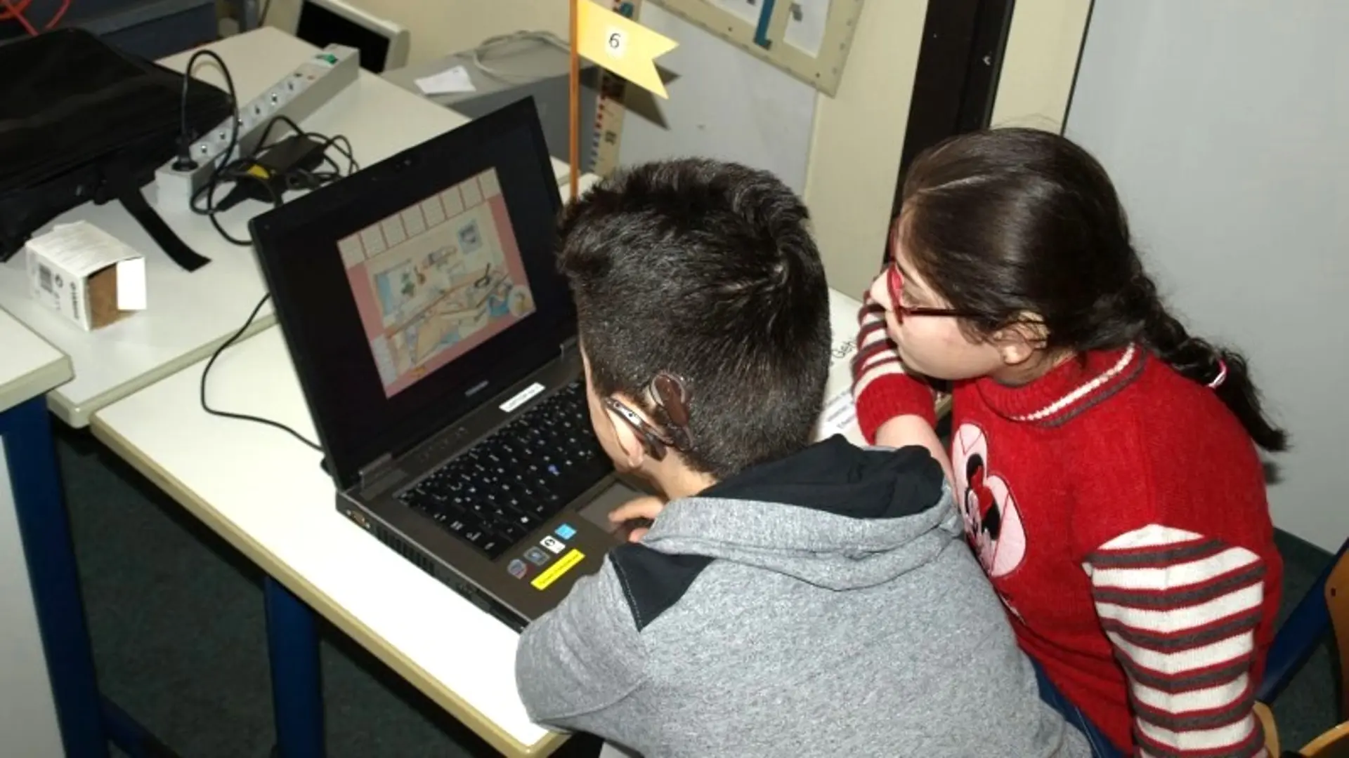 In einem Klassenraum sind zwei Kinder von hinten zu sehen. Sie schauen auf ein Notebook. Eins der Kinder trögt ein Cholear-Implantat.