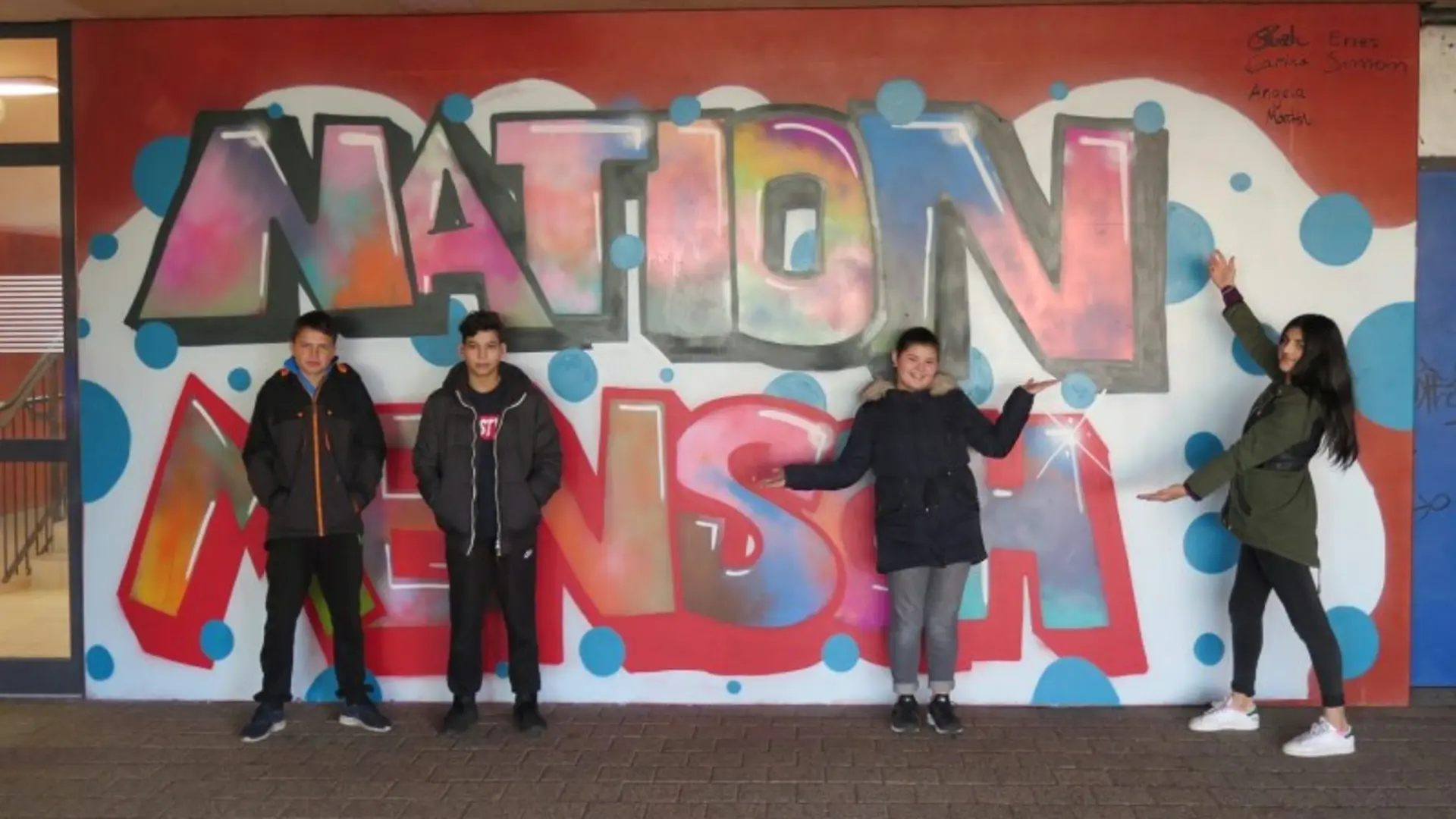 Vier Jugendliche stehen vor einem wandfüllenden bunten Graffiti mit dem Schriftzug "Nation Mensch".