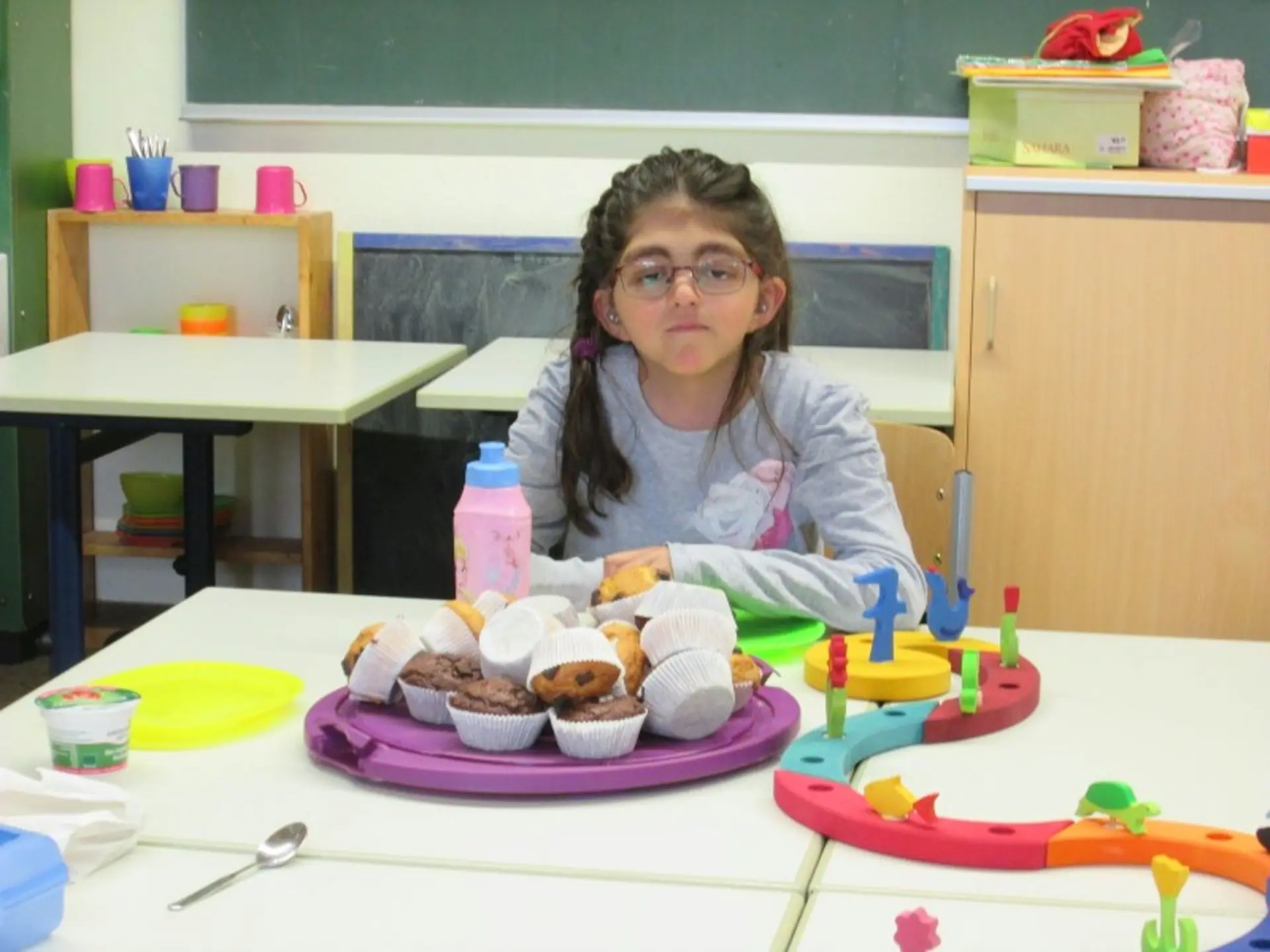 Ein Mädchen mit Brille sitz an einem Tisch. Vor ihm steht ein Teller mit vielen Muffins und ein Kerzenhalter in Form einer bunten Schlange mit der Zahl sieben.