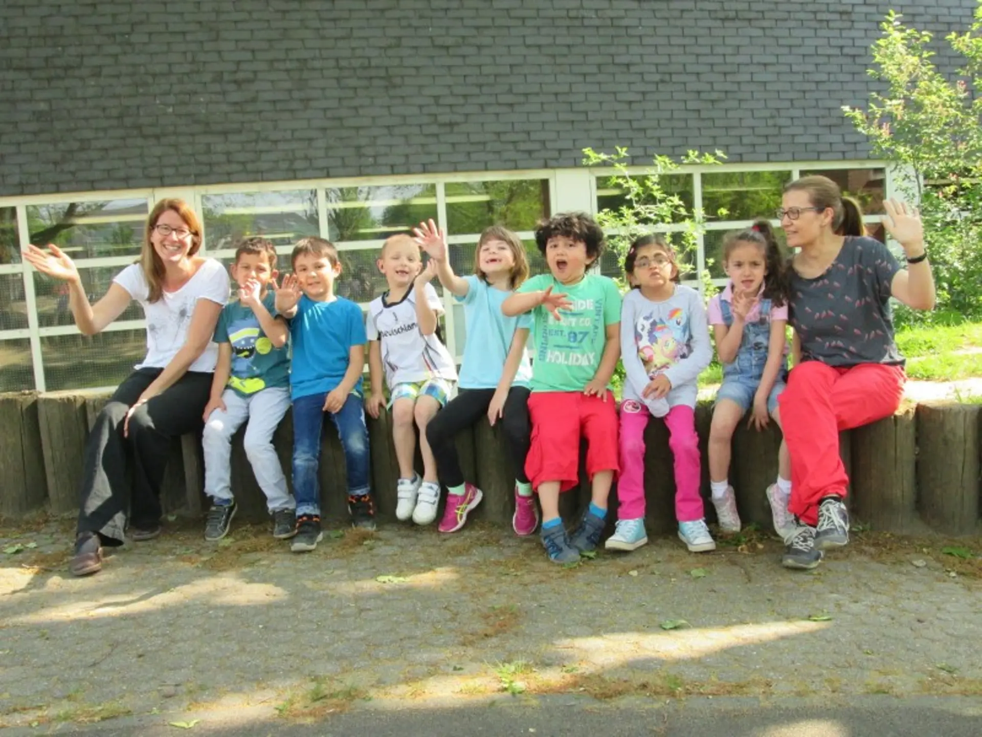 Sieben Kinder der Vorschule und zwei Lehrerinnen sitzen auf einem Mäuerchen im Freien und winken in die Kamera.