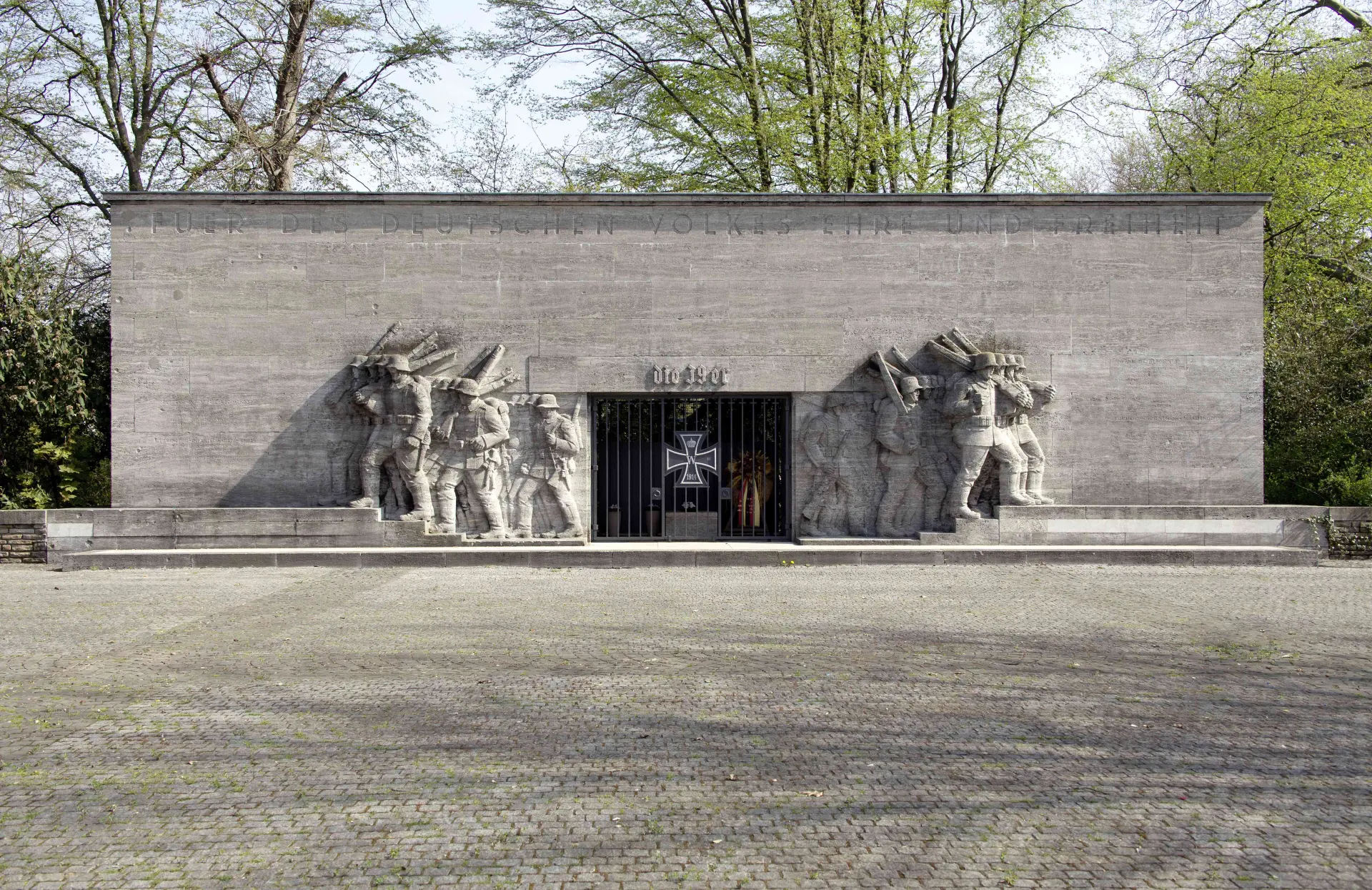 Ansicht eines nationalsozialistischen Kriegerehrenmals aus Naturstein mit reliefierten Darstellungen marschierender Soldaten in Seitansicht, vorgelagert große Platzanlage. 