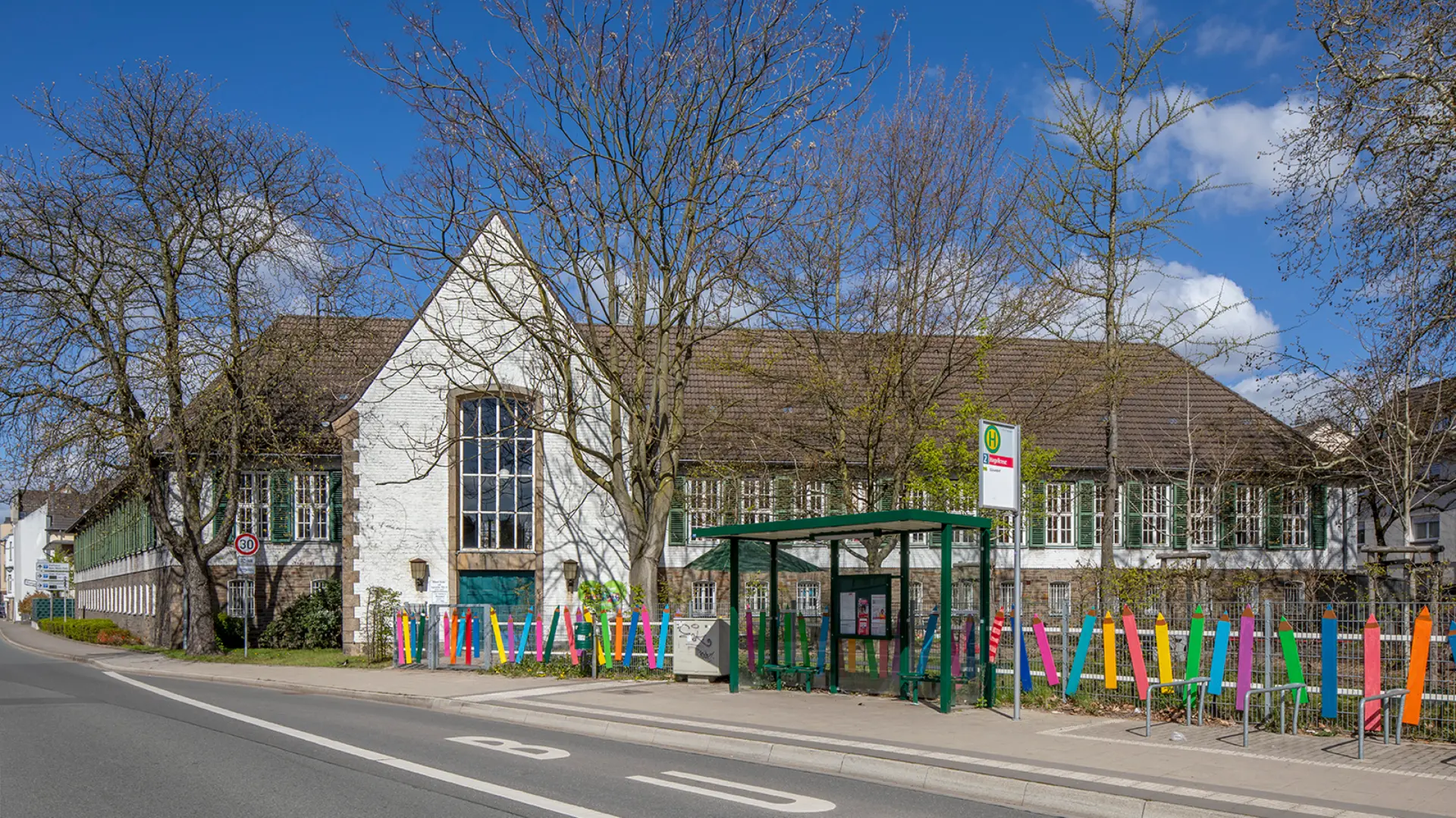 An einer Straße gelegener, winkelförmiger Ziegelbau mit weiß geschlämmter Fassade und ziegelgedecktem Walmdach, im Vordergrund Zaunanlage, die mit bunten Stiften dekoriert ist. 
