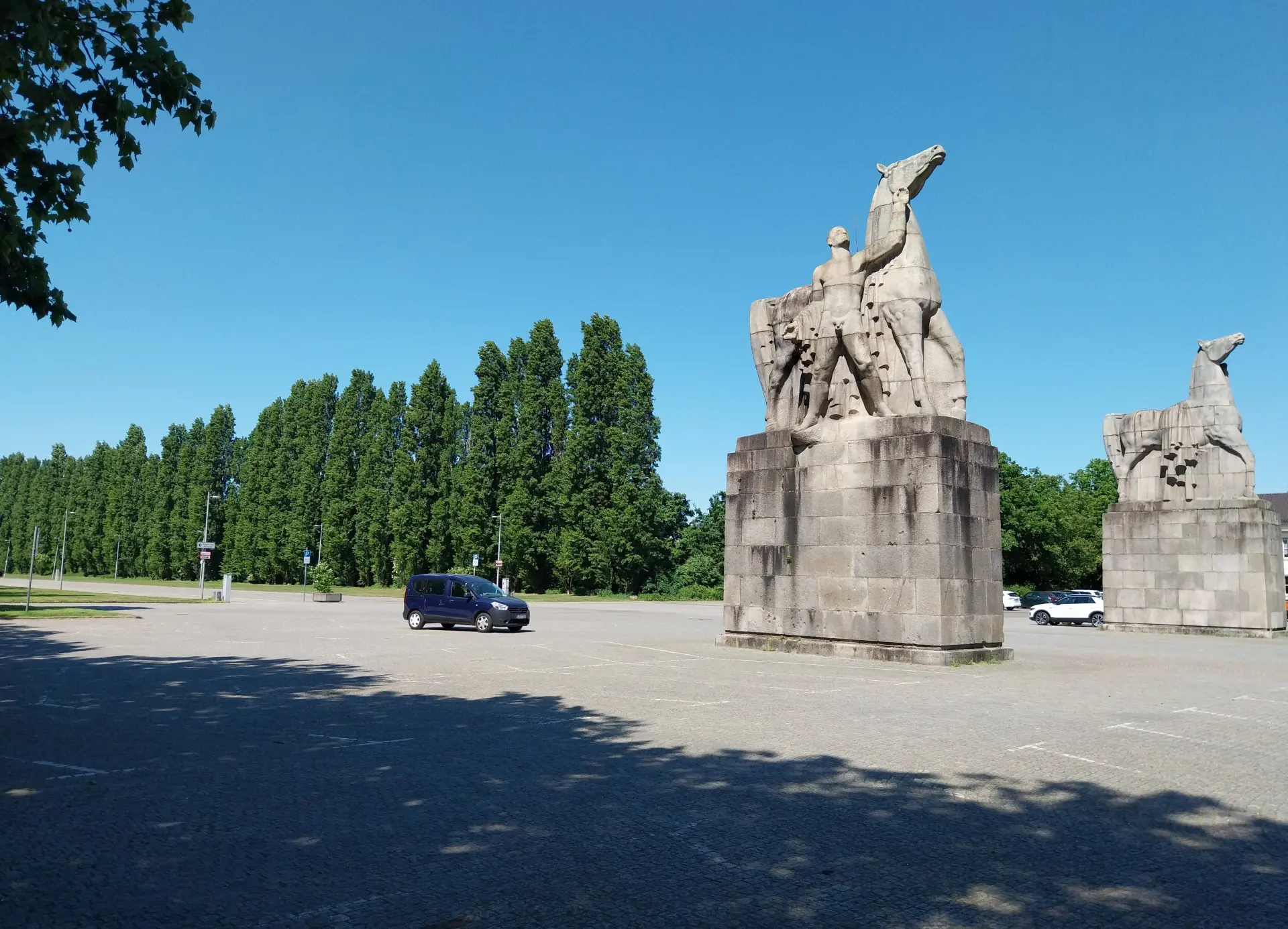 Ansicht von zwei monumentalen Pferdeskulpturen und einem Rossbändiger aus Naturstein im Eingangsbereich zum Nordpark, die vom Bildhauer Edwin Scharff anlässlich der Großen Reichsausstellung Schaffendes Volk 1937 aufgestellt wurden, im Hintergrund ist eine Pappelallee zu sehen. 