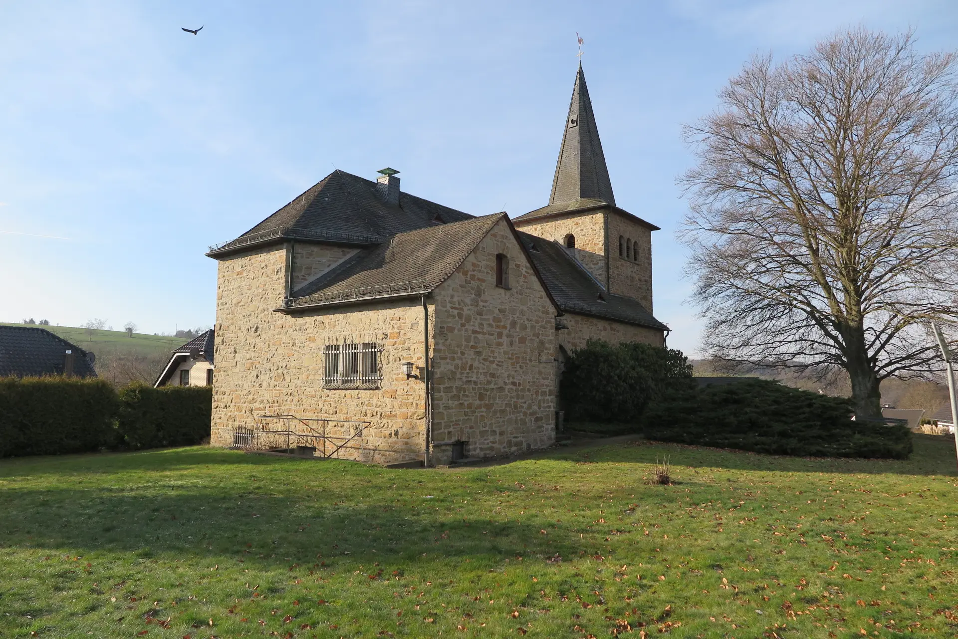 Ansicht einer Kirche aus Natursteinmauerwerk mit niedrigem Kirchturm und ziegelgedeckten Dächern, umgeben von Wiesen. 