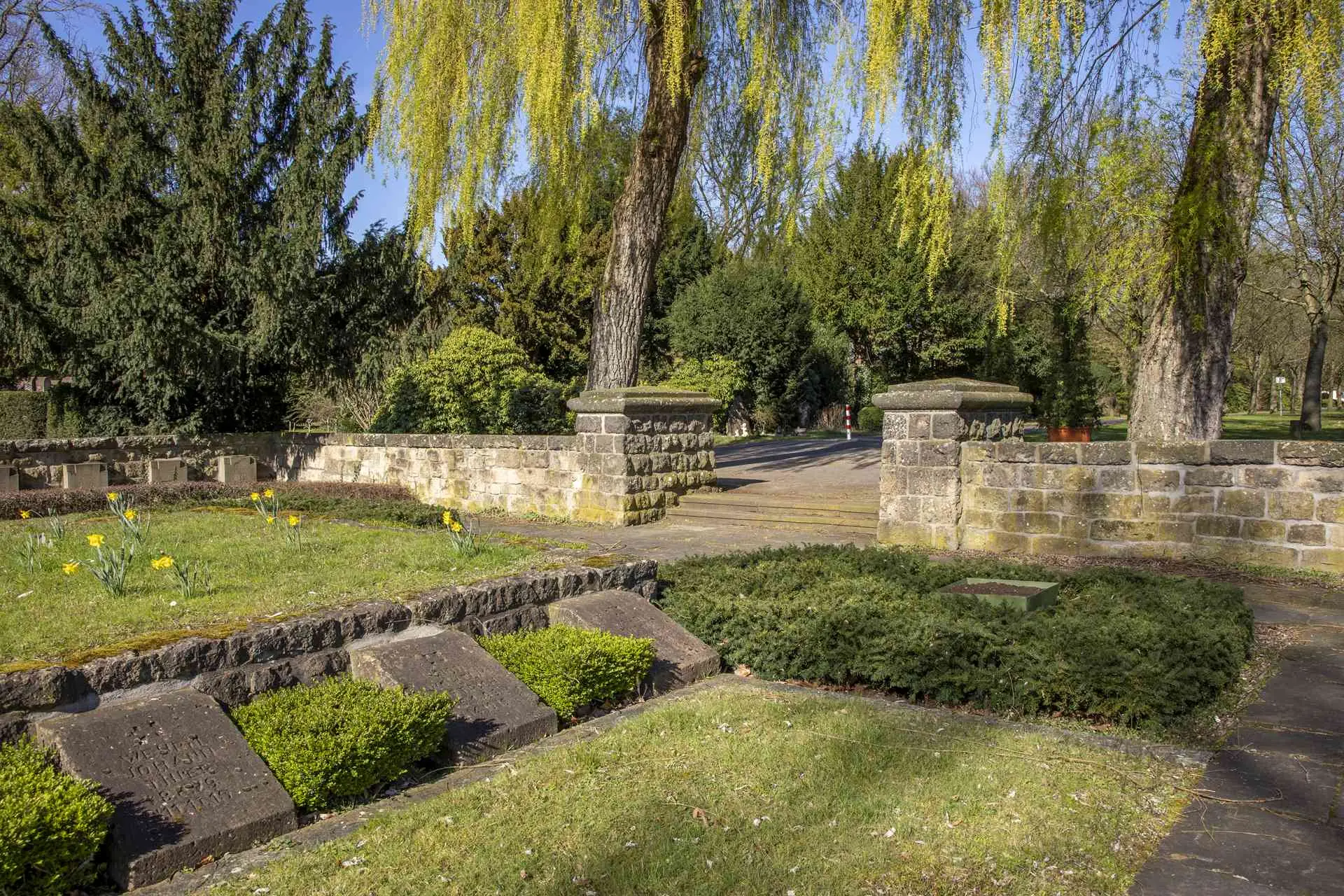 Friedhof für im Ersten Weltkrieg gefallene Soldaten mit Grabsteinen, umgeben von Hecken und Grünflächen, einer Natursteinmauer und historischem Baumbestand. 