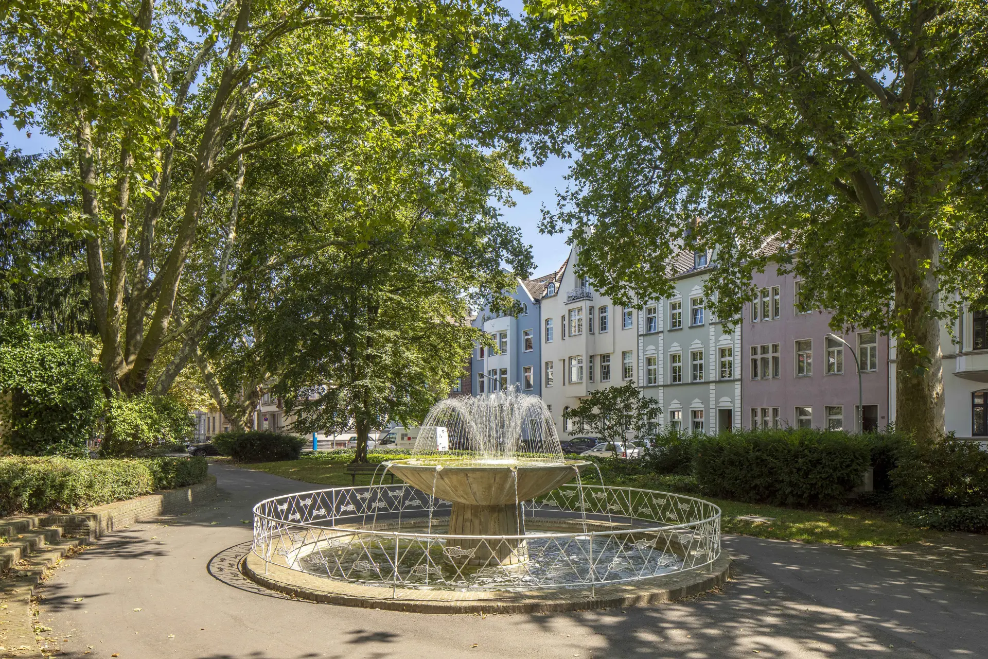 Neuss, innerstädtischer Drususplatz mit Springbrunnen, historischem Baumbestand und Wohnbebauung im Hintergrund. 