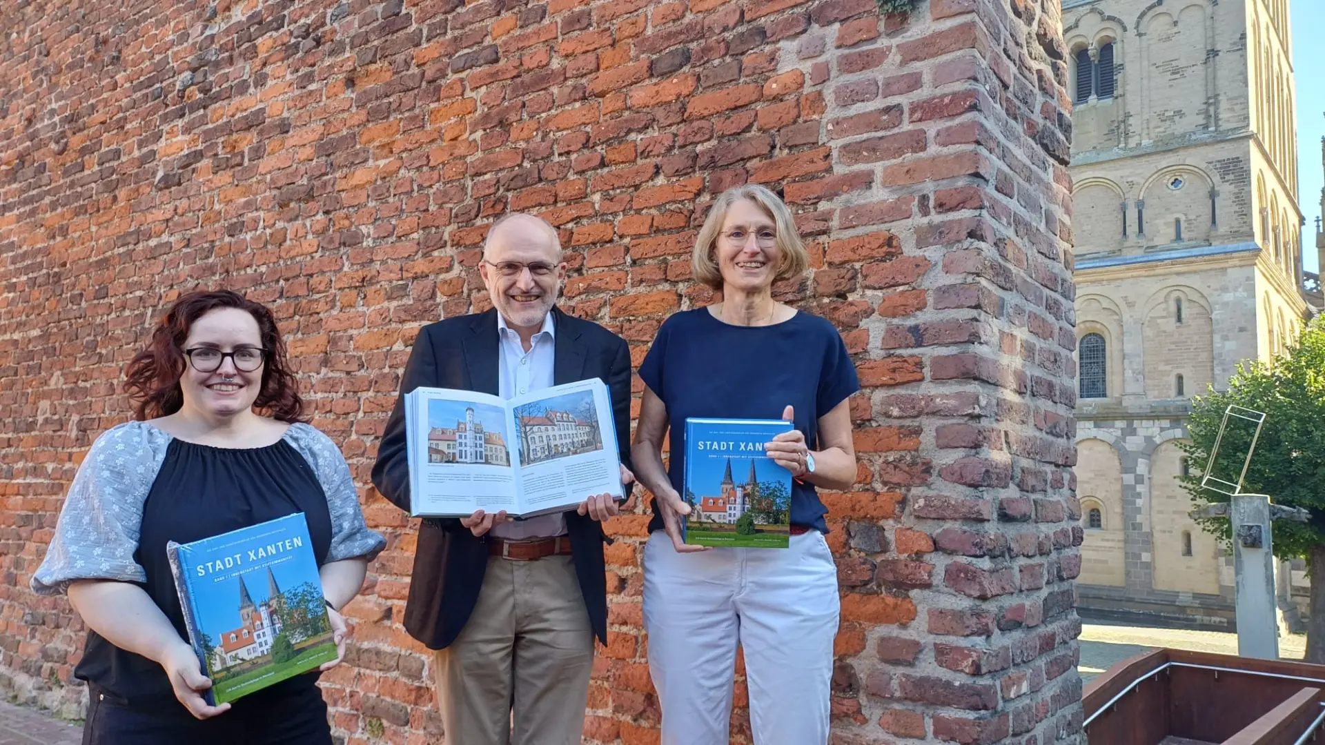 Die Fotografin, der Autor und die Landeskonservatorin vor einer Mauer mit dem Xantener Dom im Hintergrund halten das neue Buch in den Händen.