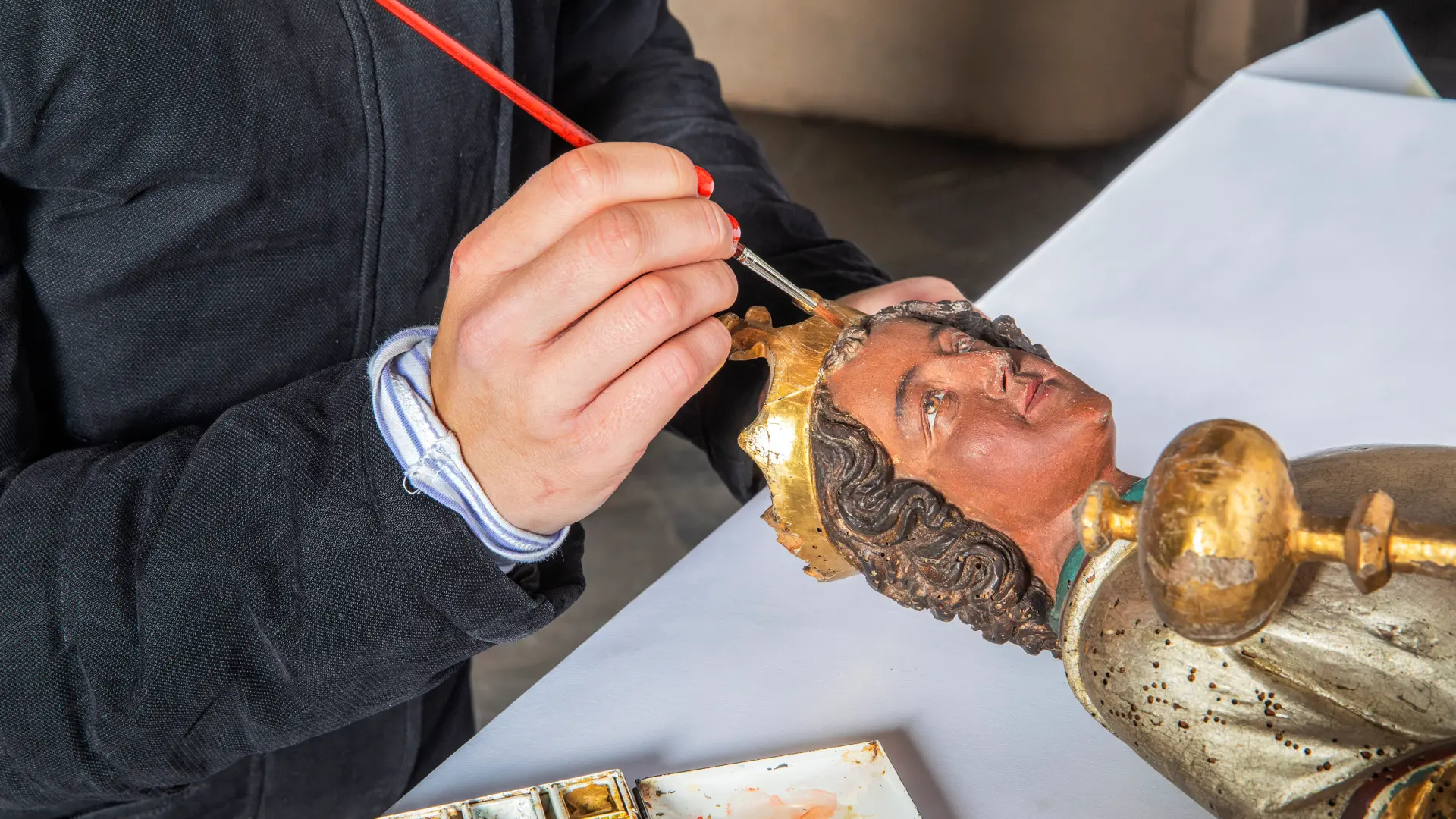 Mit dem Pinsel wird eine Farbretusche am Kopf einer hölzernen Figur ausgeführt. 