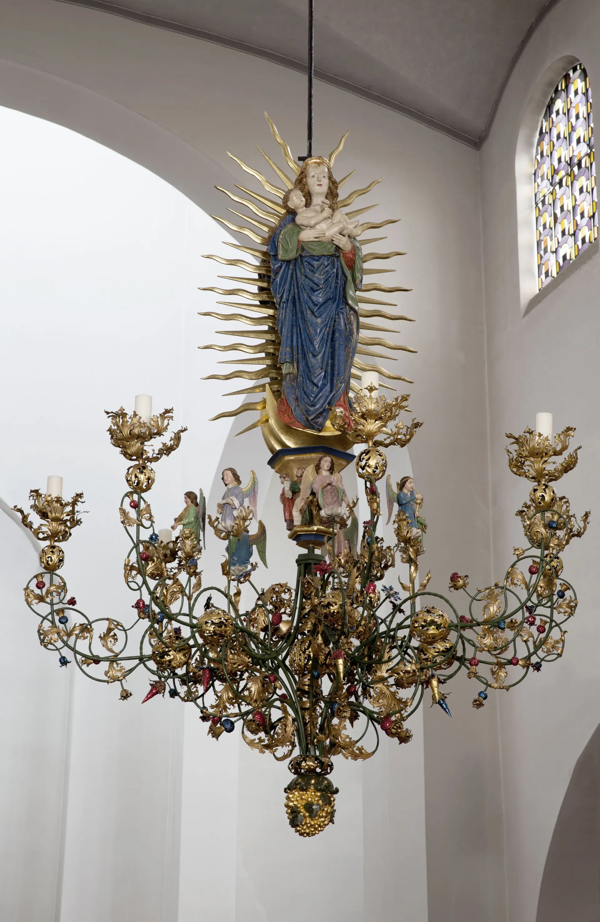 Ein 337 cm hoher und 270 cm breiter Metallleuchter mit sieben aus Rankenwerk bestehenden Leuchterarmen und einer zentralen, doppelten Marienfigur, die ein Strahlenkranz umfasst.