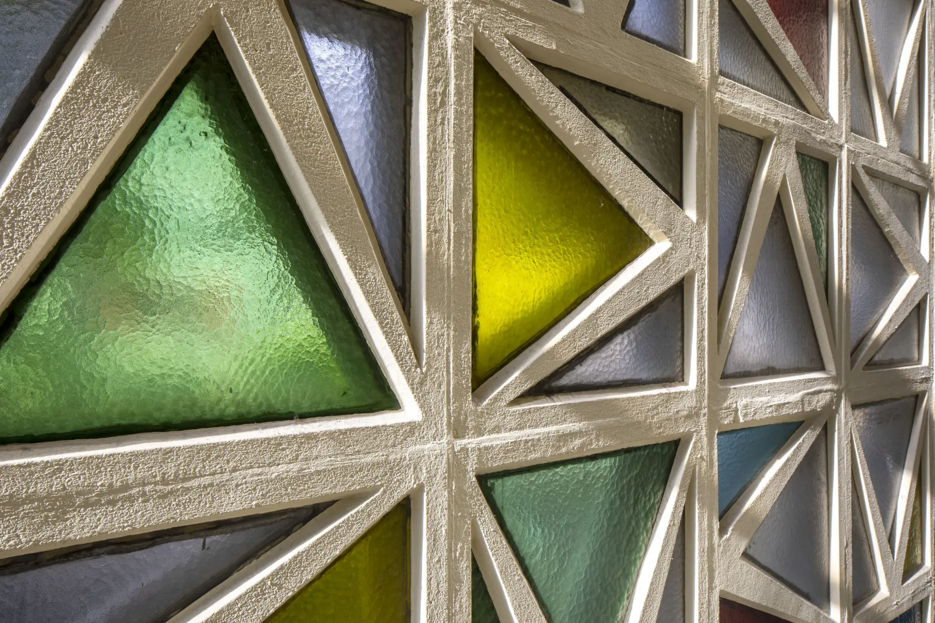 Buntverglasung in einer gitterartigen Struktur aus quadratischen, filigranen Betonrahmen mit einer abwechselnd stehenden oder liegenden dachförmigen Unterteilung, die die Glasfläche in drei  Dreiecke unterschielicher Größe unterteilt. 