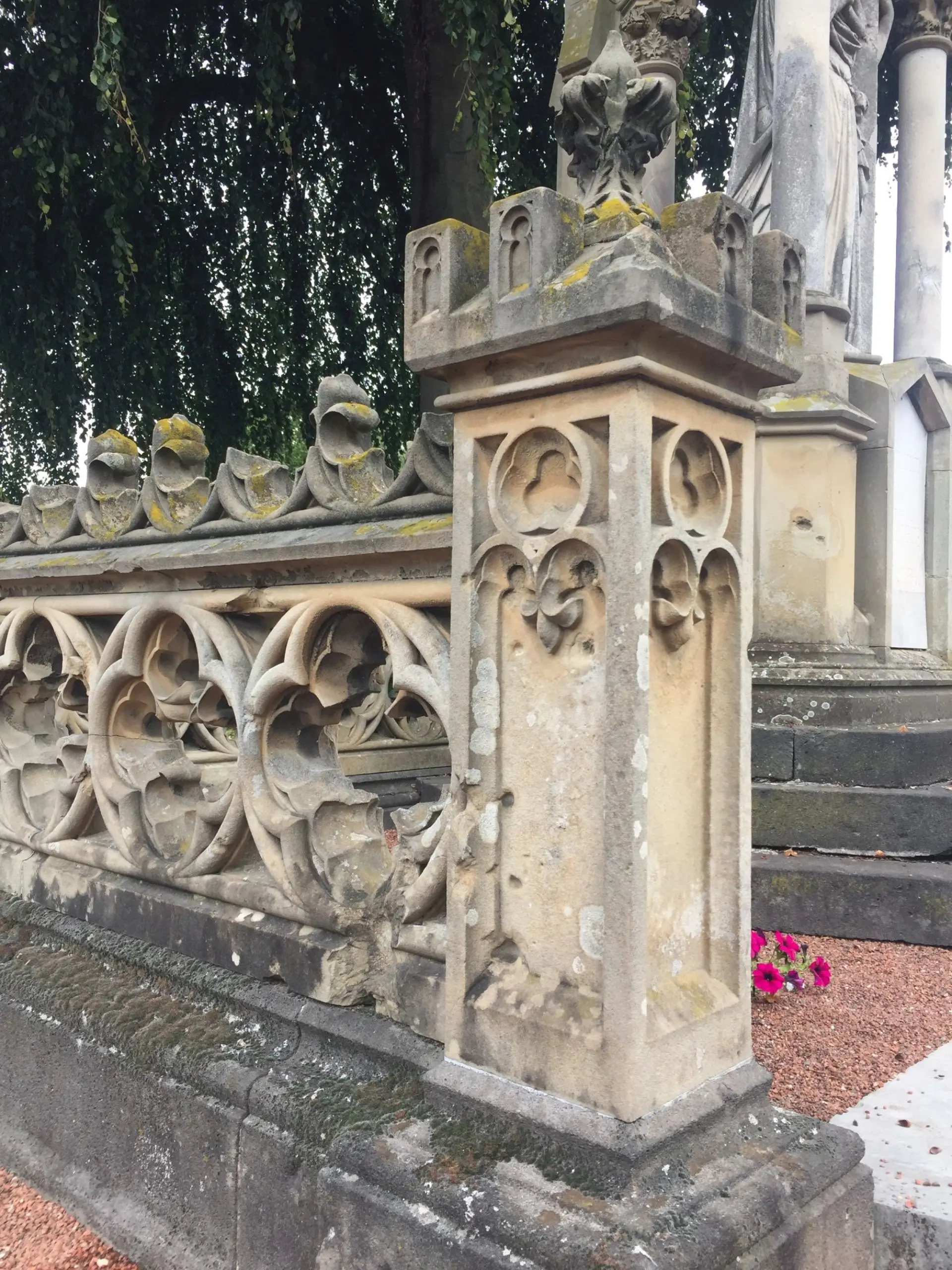 Brüstungselement des Grabmals Lensing auf dem Xantener Friedhof aus hellbraunem Sandstein mit Verschmutzungen und Flechten, die die Oberfläche besiedeln sowie Schäden an der Steinsubstanz durch Kriegseinwirkungen und Vandalismus am Maßwerkkamm.